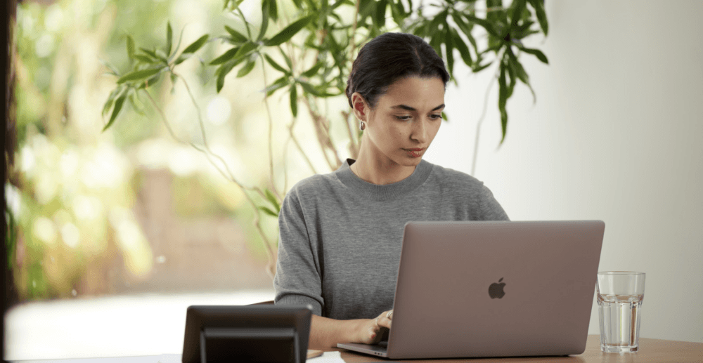 Woman working on a laptop at a wooden table with a glass of water, surrounded by greenery in a bright indoor setting.