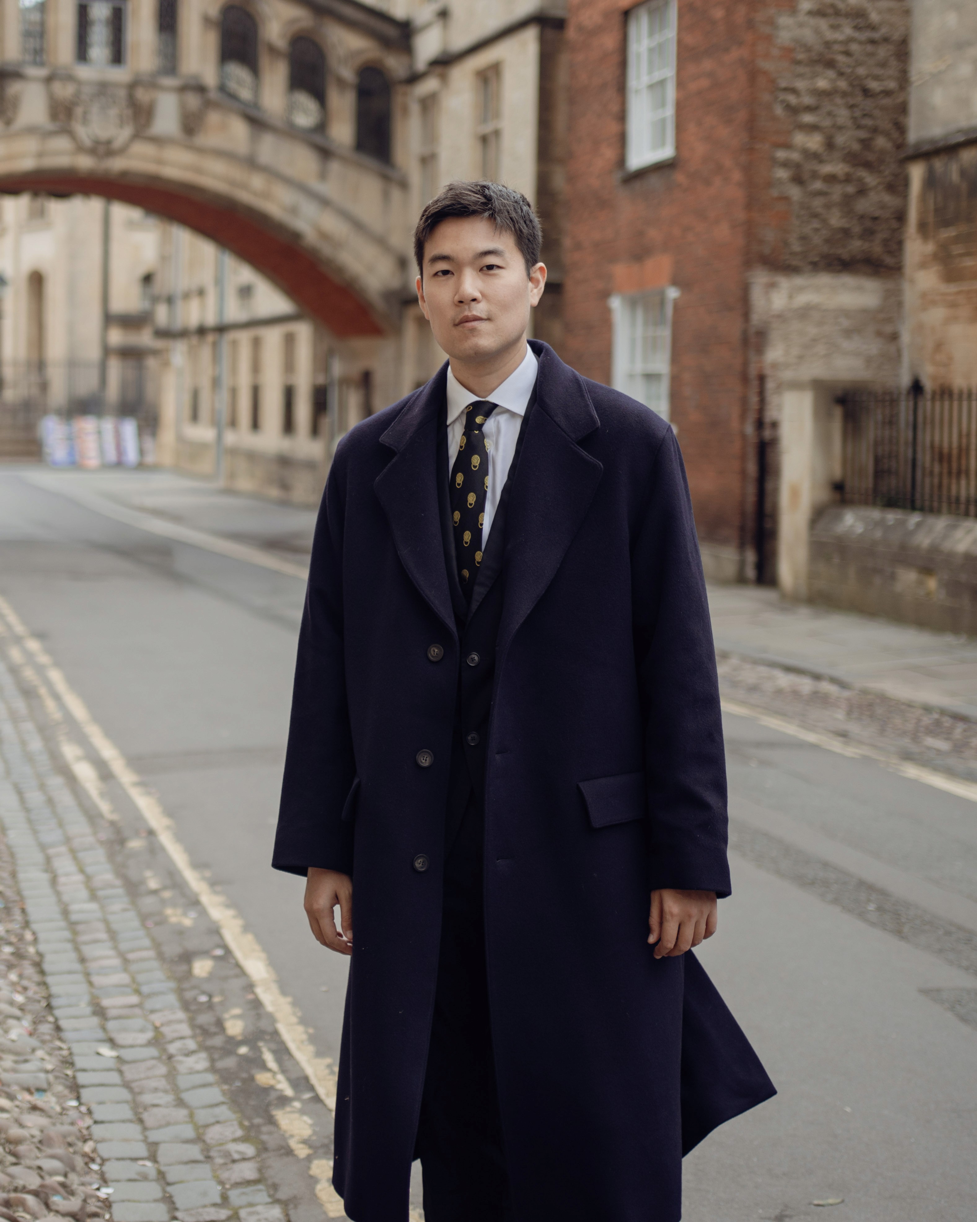 A smiling man in a dark shirt and jeans sits casually on a stool against a dark grey background, looking off to the side.