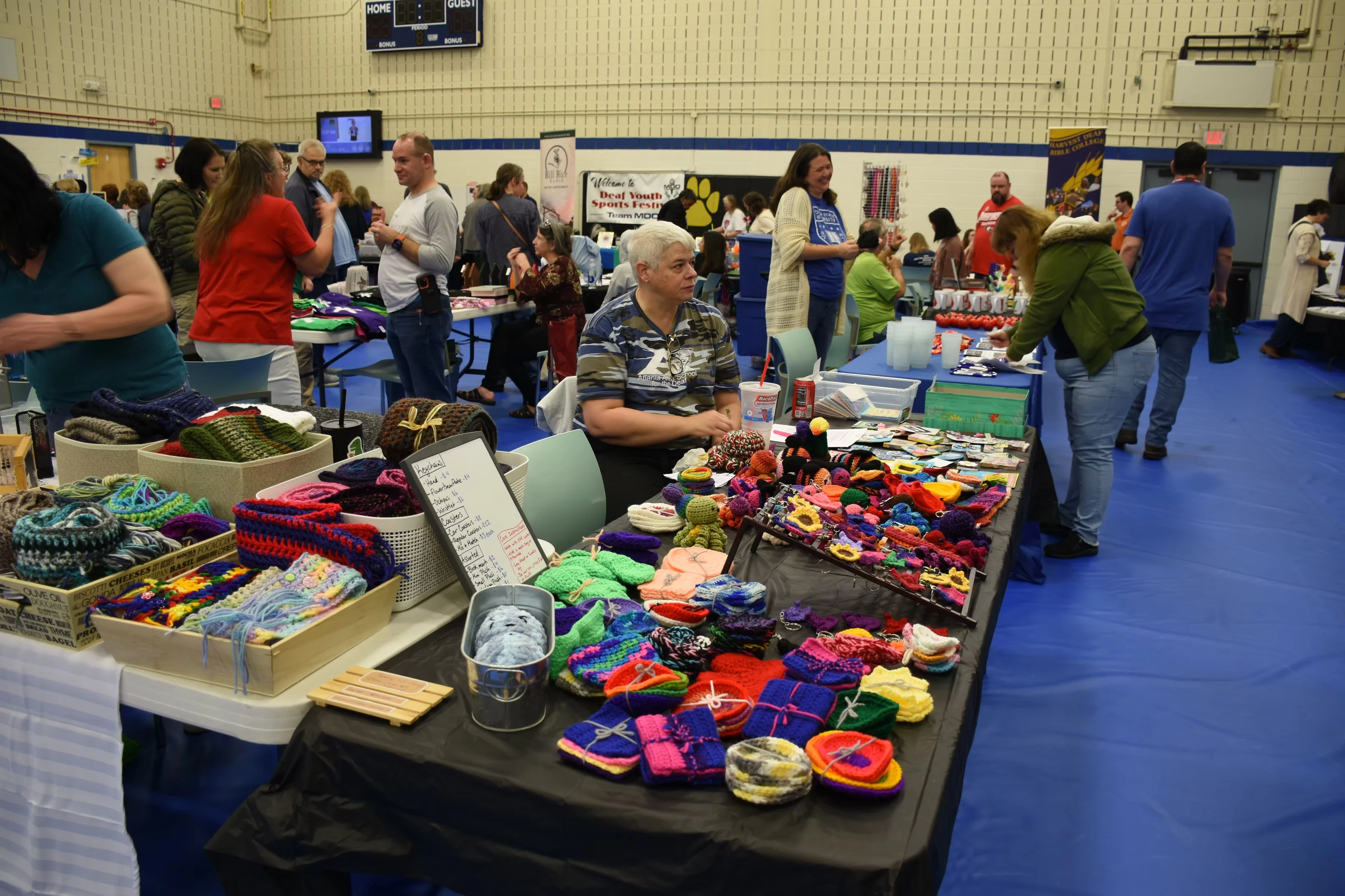 A vendor sits behind a table at a craft fair, displaying colorful handmade knitted and crocheted items such as hats, gloves, and scarves.