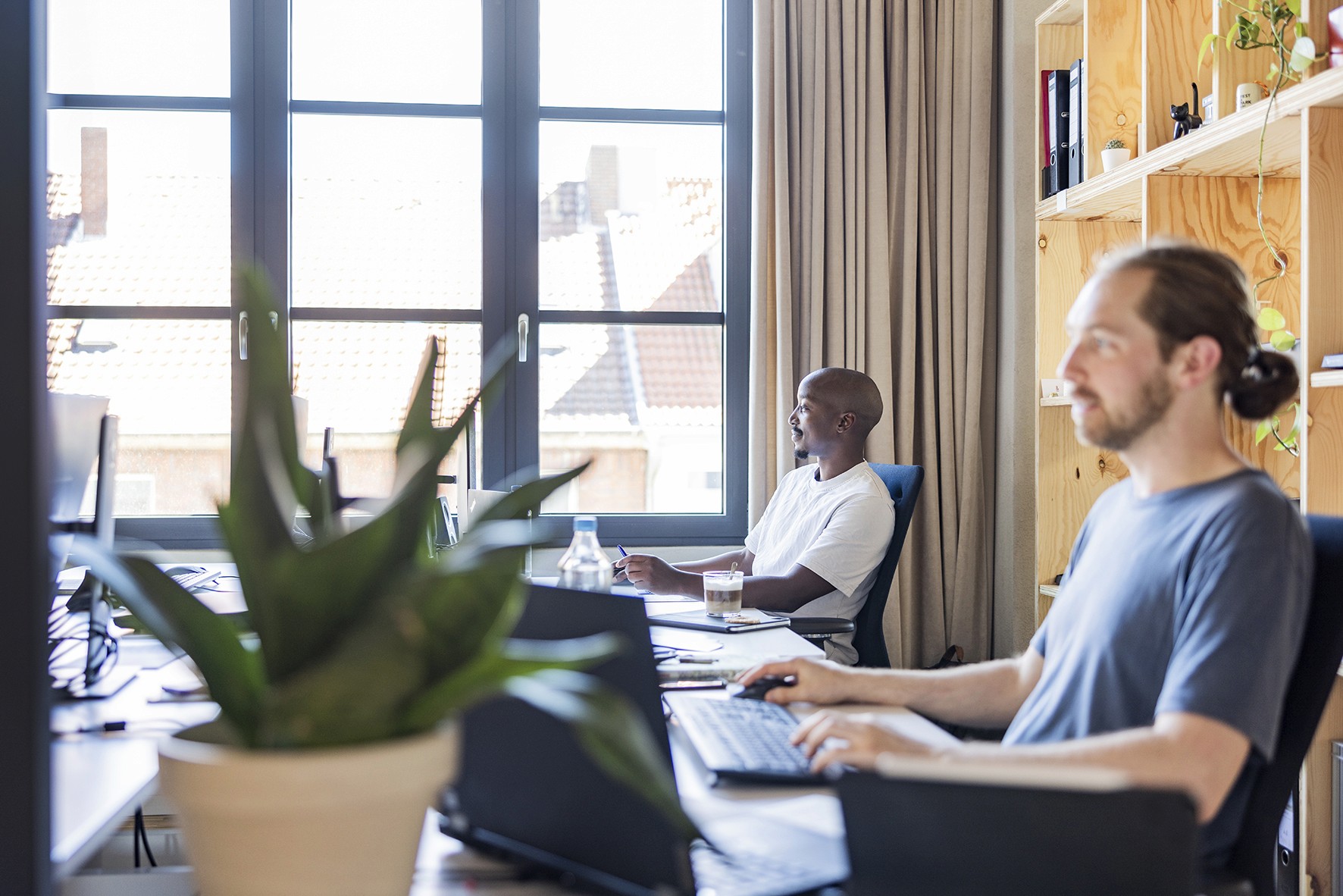 Zwei Personen arbeiten an Schreibtischen in einem hellen Büro mit großen Fenstern, Pflanzen und Holzregalen; moderner Arbeitsraum von MAAS & PARTNER.