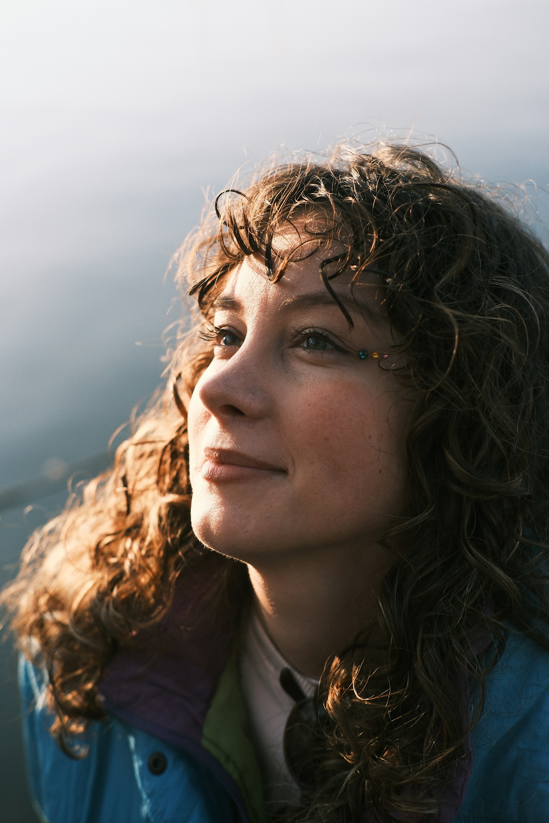 A close-up portrait of a woman with curly brown hair looking upward. She has small, colorful decorative gems near her eye and is lit by warm, low-angle sunlight.