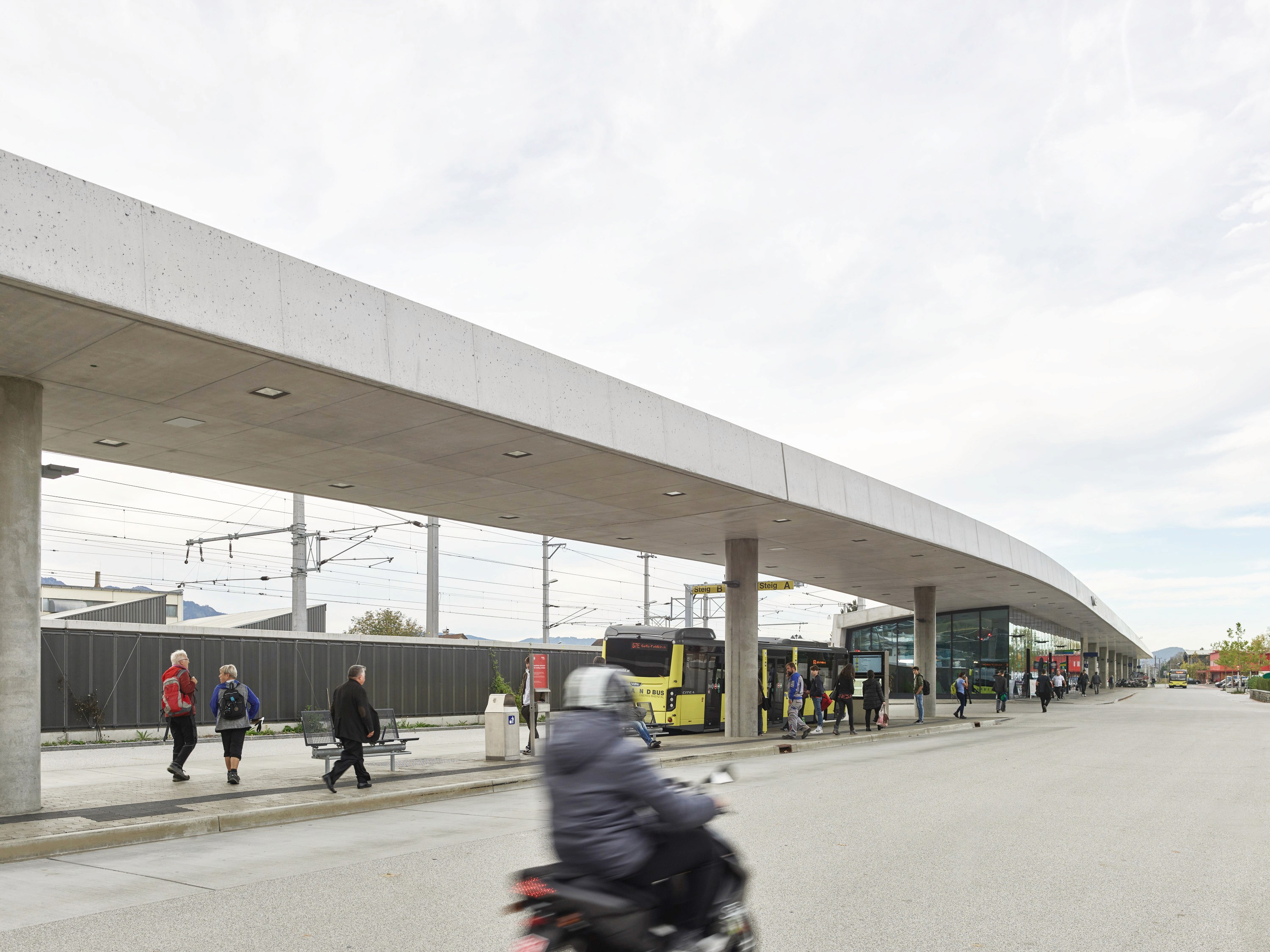 Blick auf den Bahnhof Rankweil mit lang gezogenem Beton-Vordach, mehreren wartenden Menschen und einem gelben Bus an der Haltestelle und Mopedfahrer im Vordergrund.
