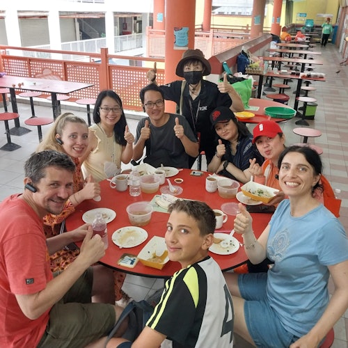 Eight people smiling and giving thumbs up while seated around a table with food in a casual dining area with red tables and chairs.