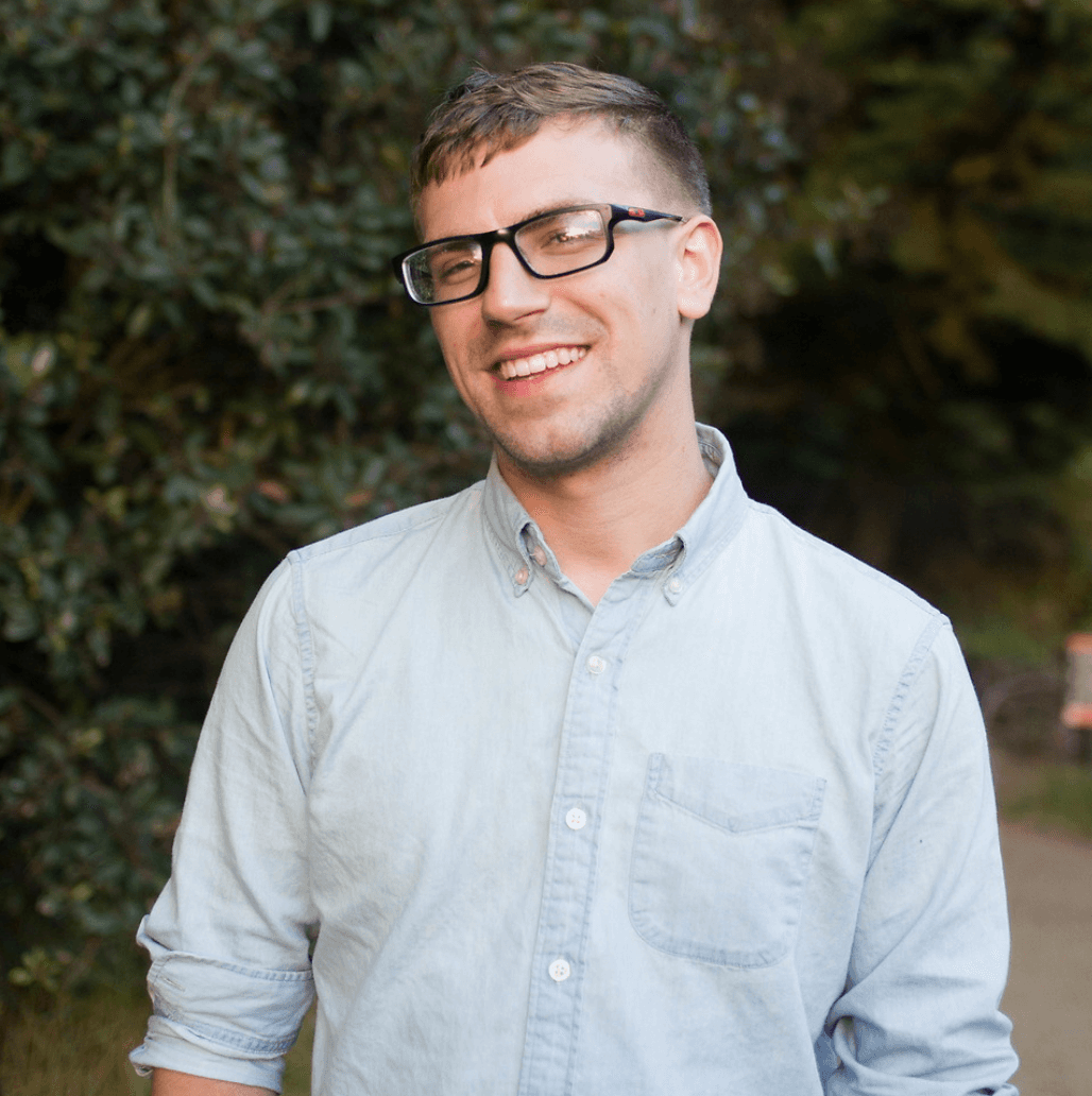 A person in glasses wearing a light blue button-up shirt is outdoors, smiling in front of a leafy green background.