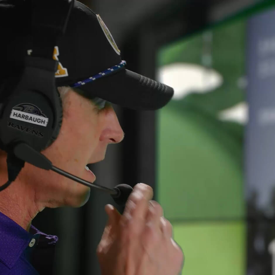 A football coach wearing a headset and cap speaks into a microphone while focusing on a screen.