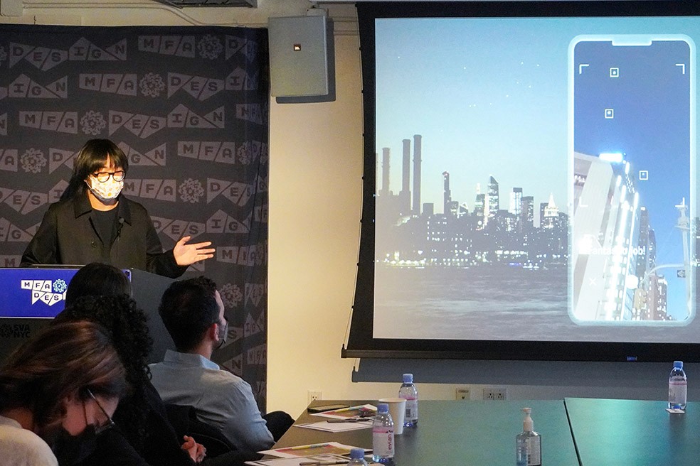 A man is giving a speech in front of other people, and on the right side is an image projected with a phone screen with a cityscape image on it.