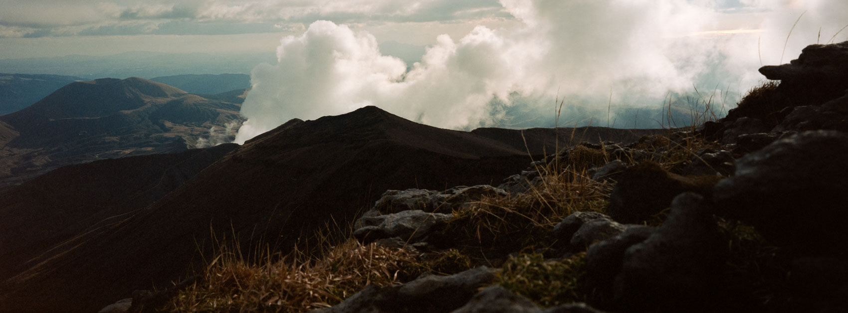 Japan hiking at Mount Aso: step from the volcano appear in the back behind some mountain ridges, sunny landscape with rocks on the ground and clouds in the sky, captured on Kodak Portra film in Kyushu's otherworldly terrain.