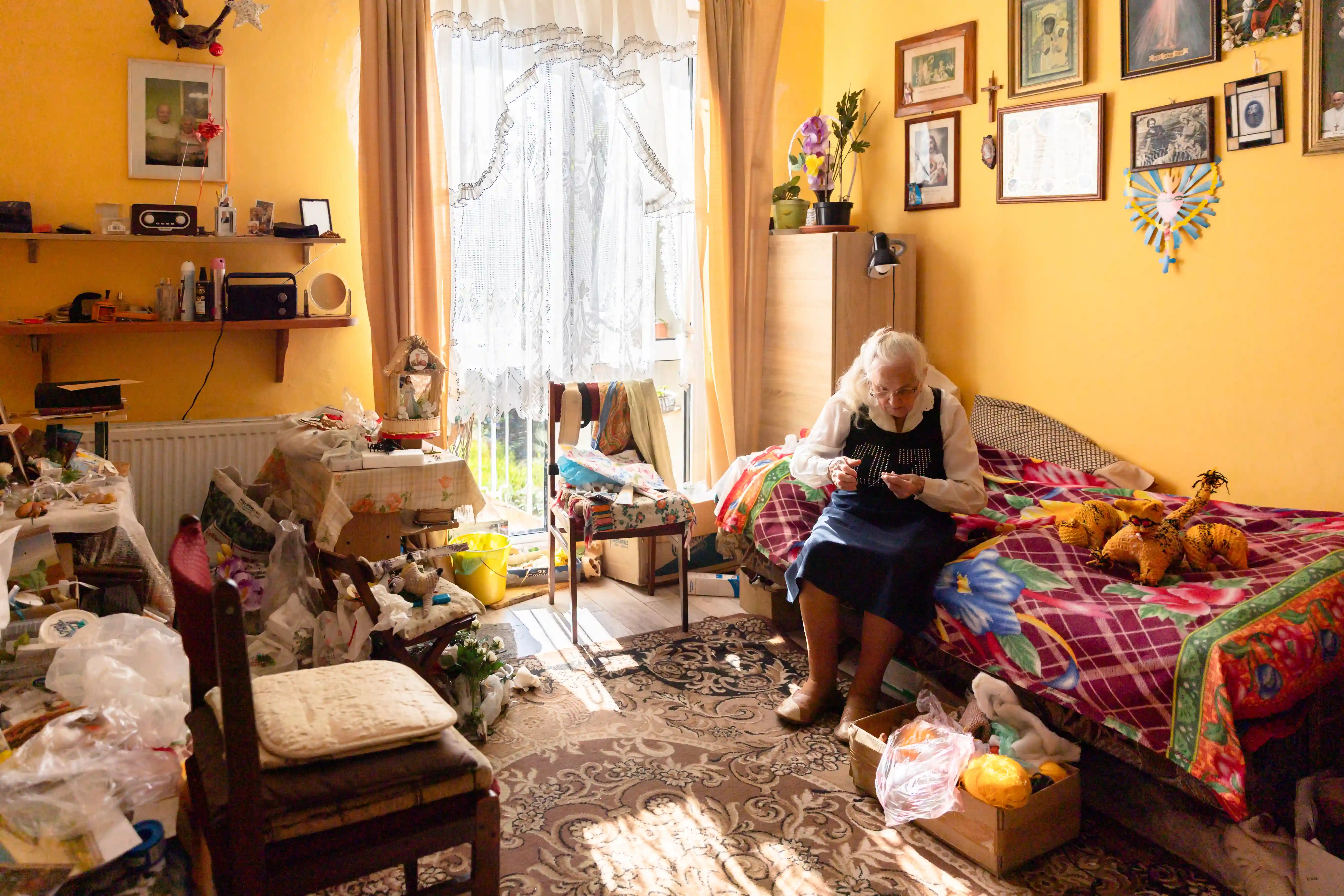 Starsza pani siedzi na łóżku w słonecznym pokoju. Wokół znajduje się wiele przedmiotów, na ścianie wiszą obrazy.