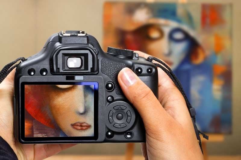 Hands holding a camera focused on an abstract painting of a face.