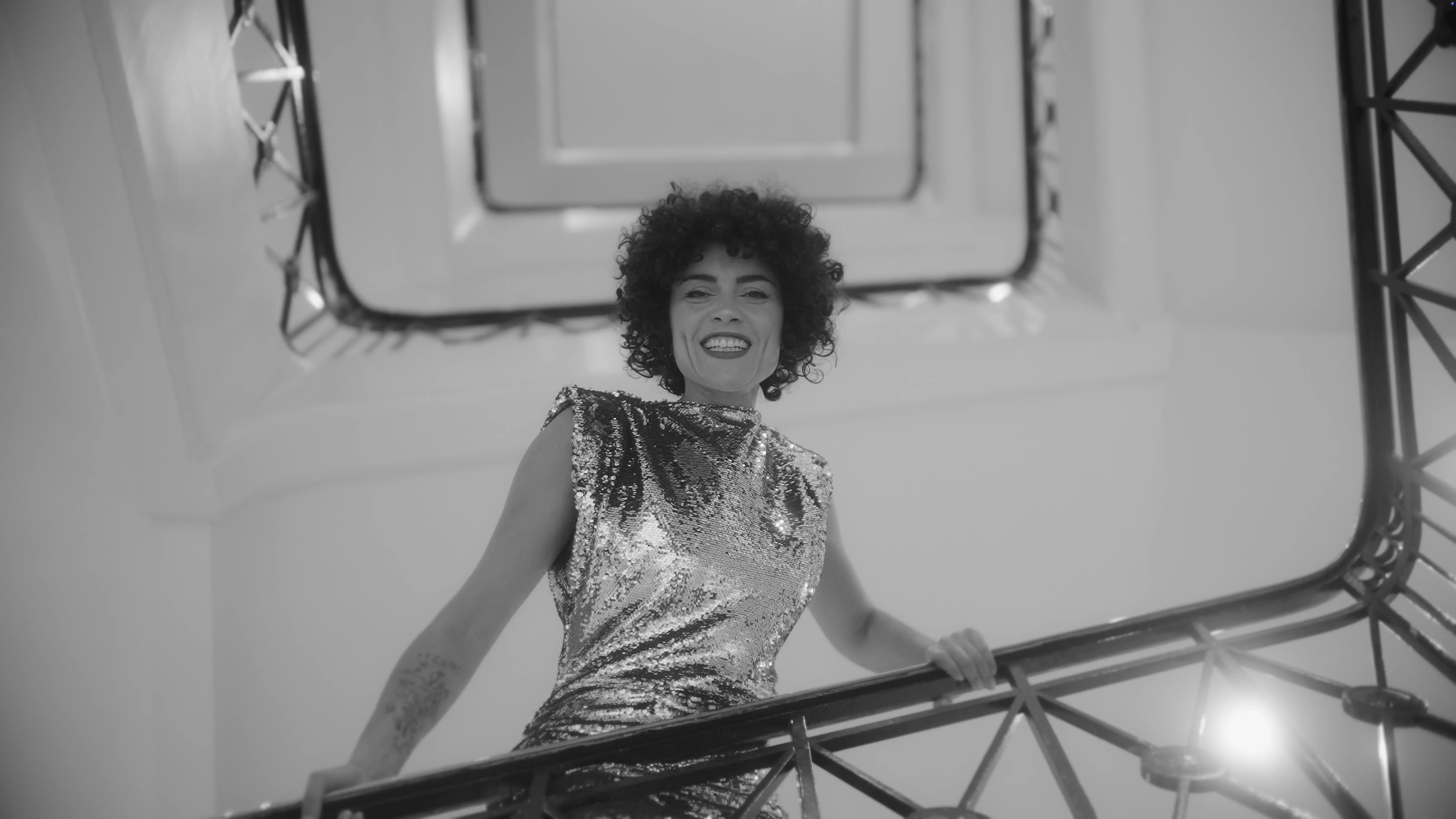 A person in a shimmering sequin dress stands confidently on a staircase, surrounded by elegant architectural details in a monochrome setting.