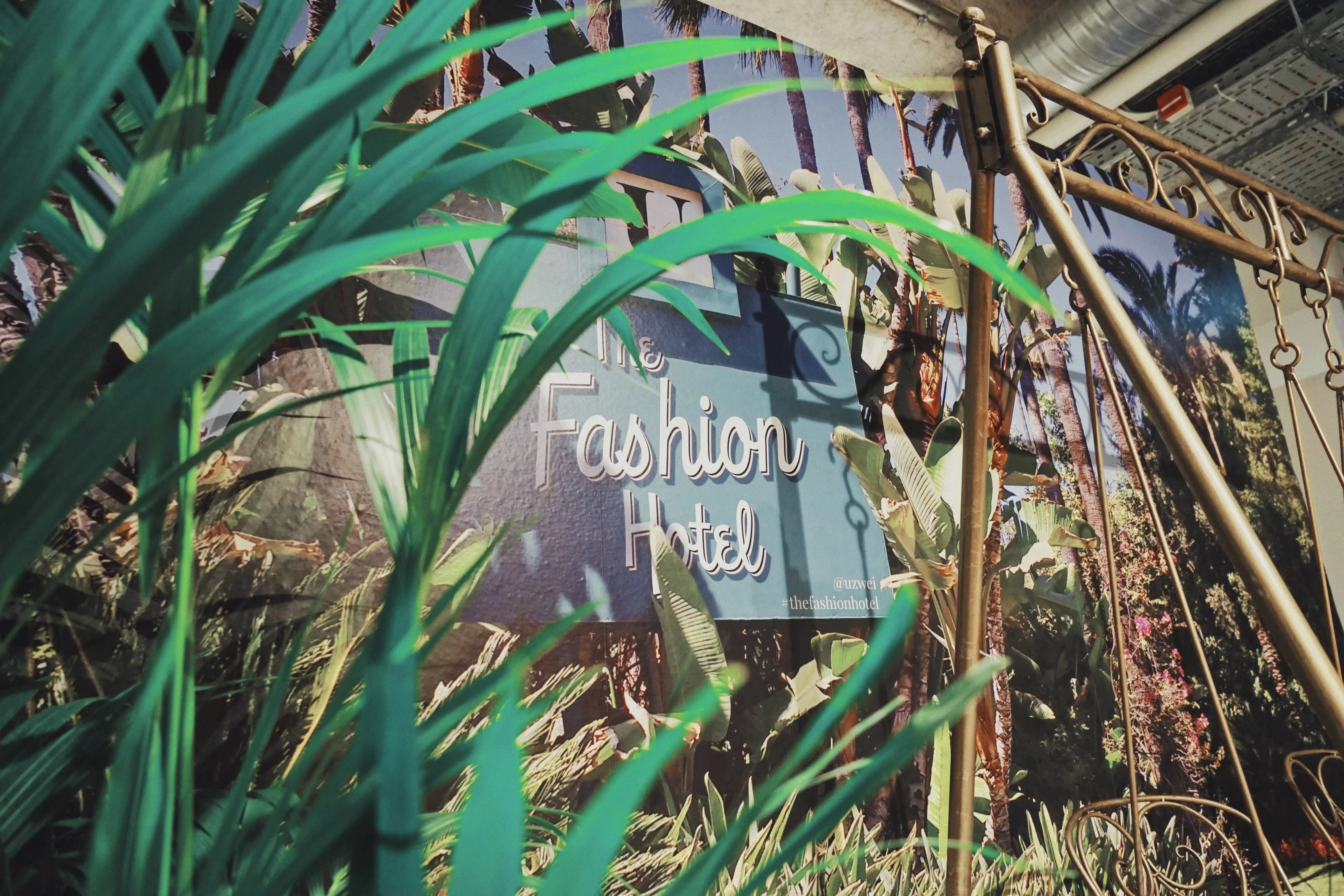 Retro neon-style The Fashion Hotel sign printed on a palm-filled mural at UZWEI, seen through green artificial palm leaves with a golden swing frame.