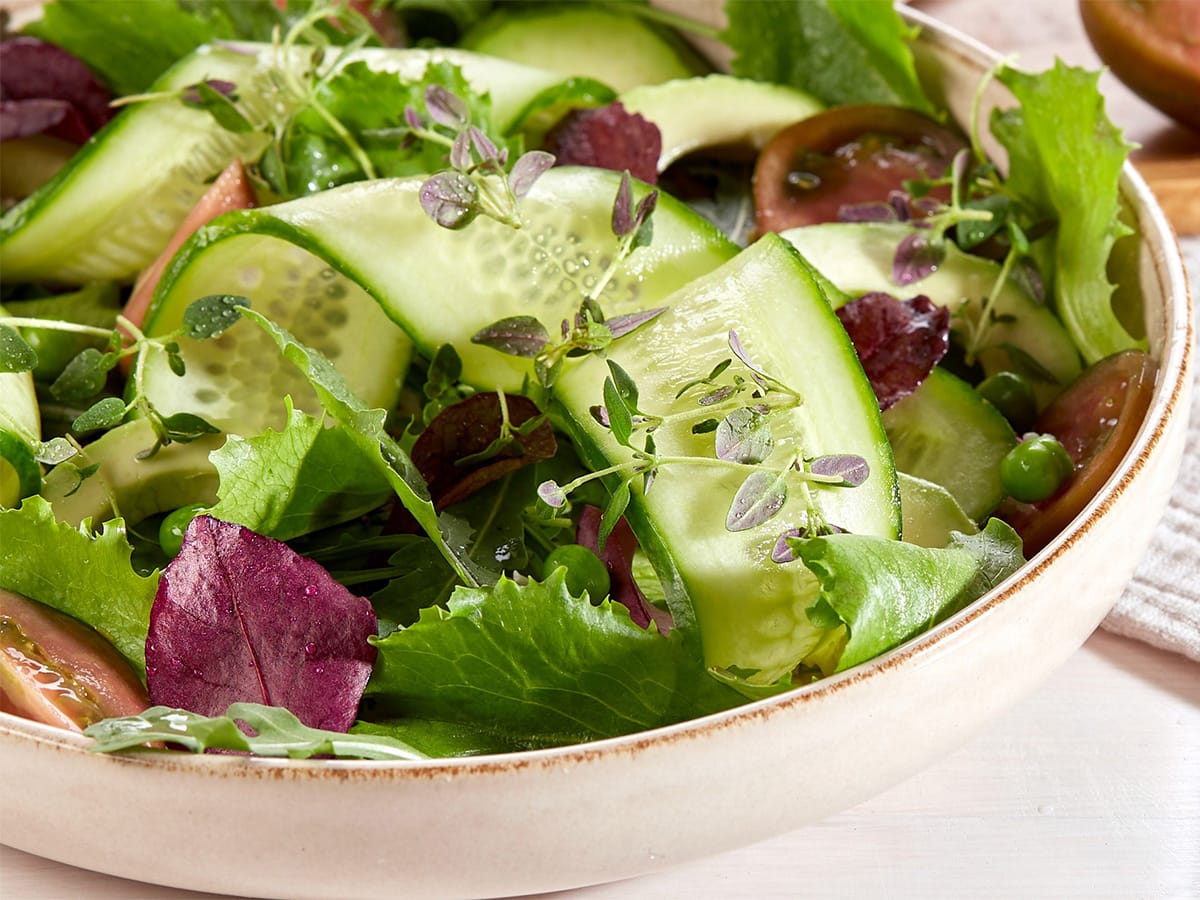 A fresh garden salad in a ceramic bowl features crisp cucumber slices, vibrant green leafy lettuce, ripe tomato wedges, and a sprinkle of delicate herbs, set on a light-colored table with a wooden spoon nearby.