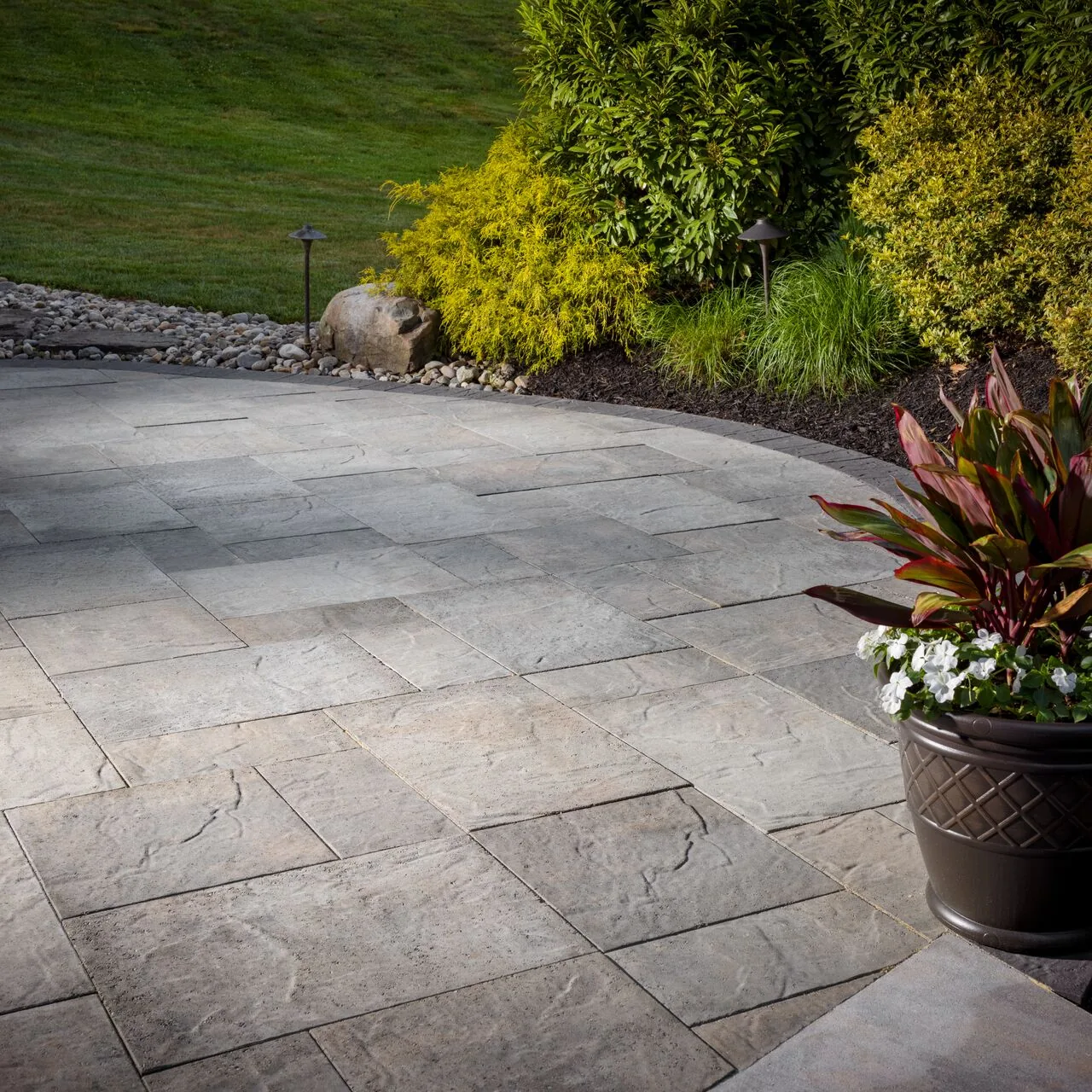 Large rectangular paving with a curved border beside shrubs and a planted pot.
