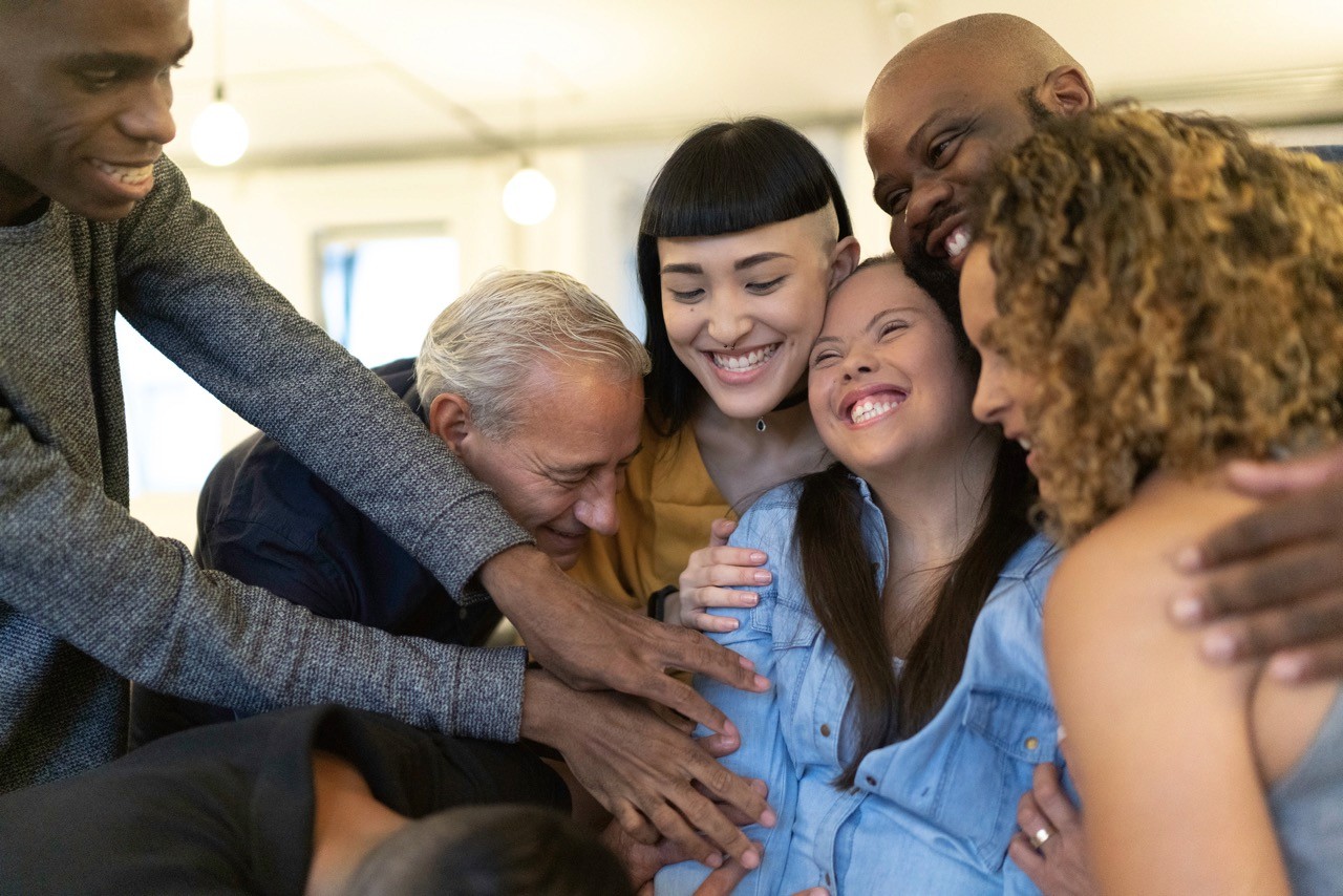 A joyful group of people, smiling and embracing a woman with a visible baby bump, gather closely and touch her belly in celebration. The scene radiates happiness and togetherness in a bright indoor setting.