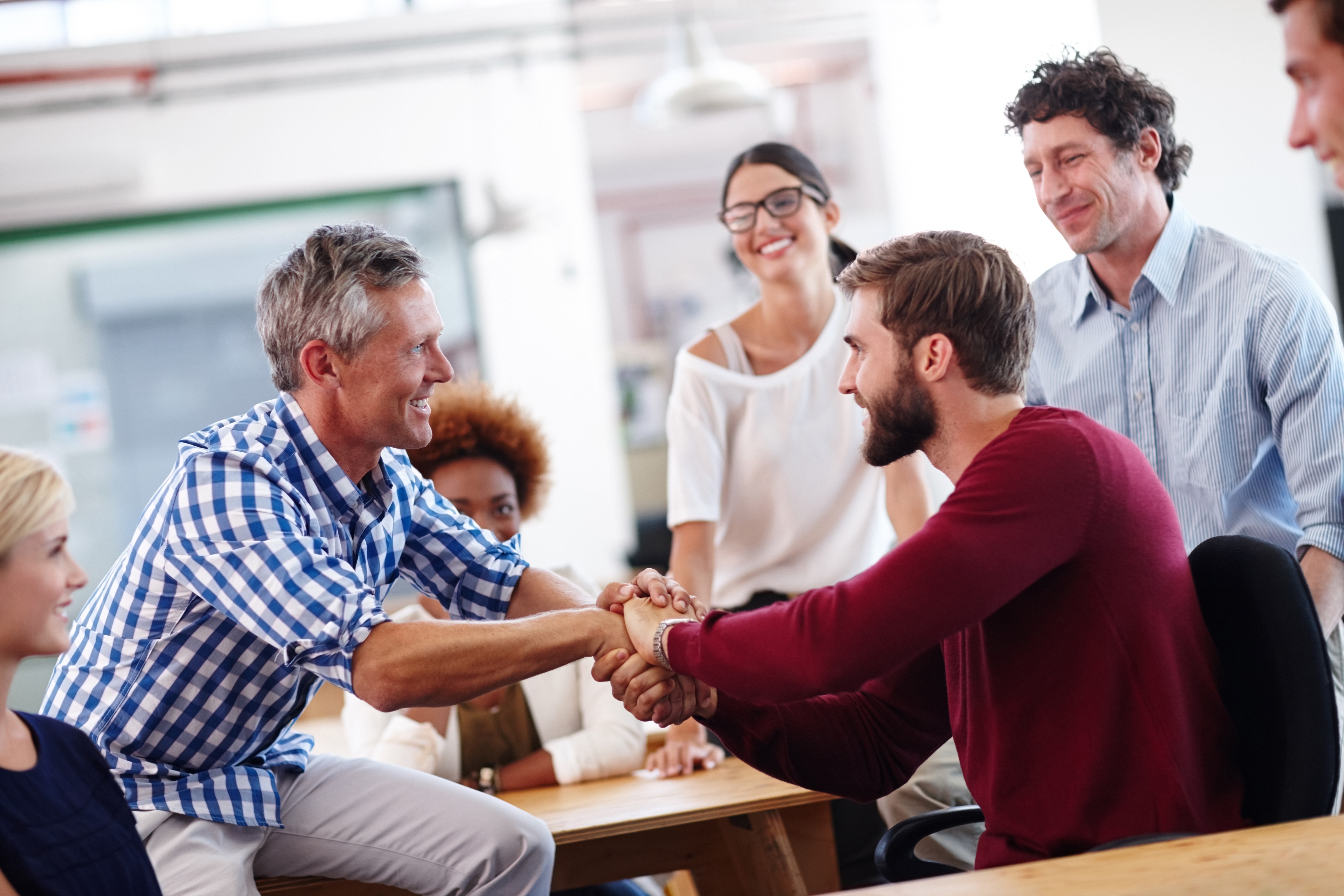 Two colleagues smiling while working together at a desk, one wearing a cream jacket and the other a beige cap and glasses.