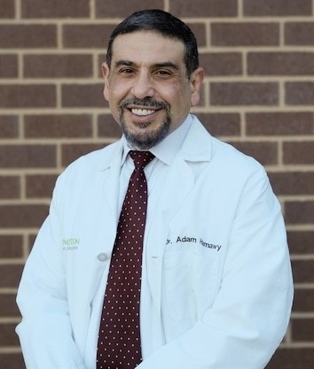 A smiling man in a light blue dress shirt and maroon tie stands in front of a brick wall.