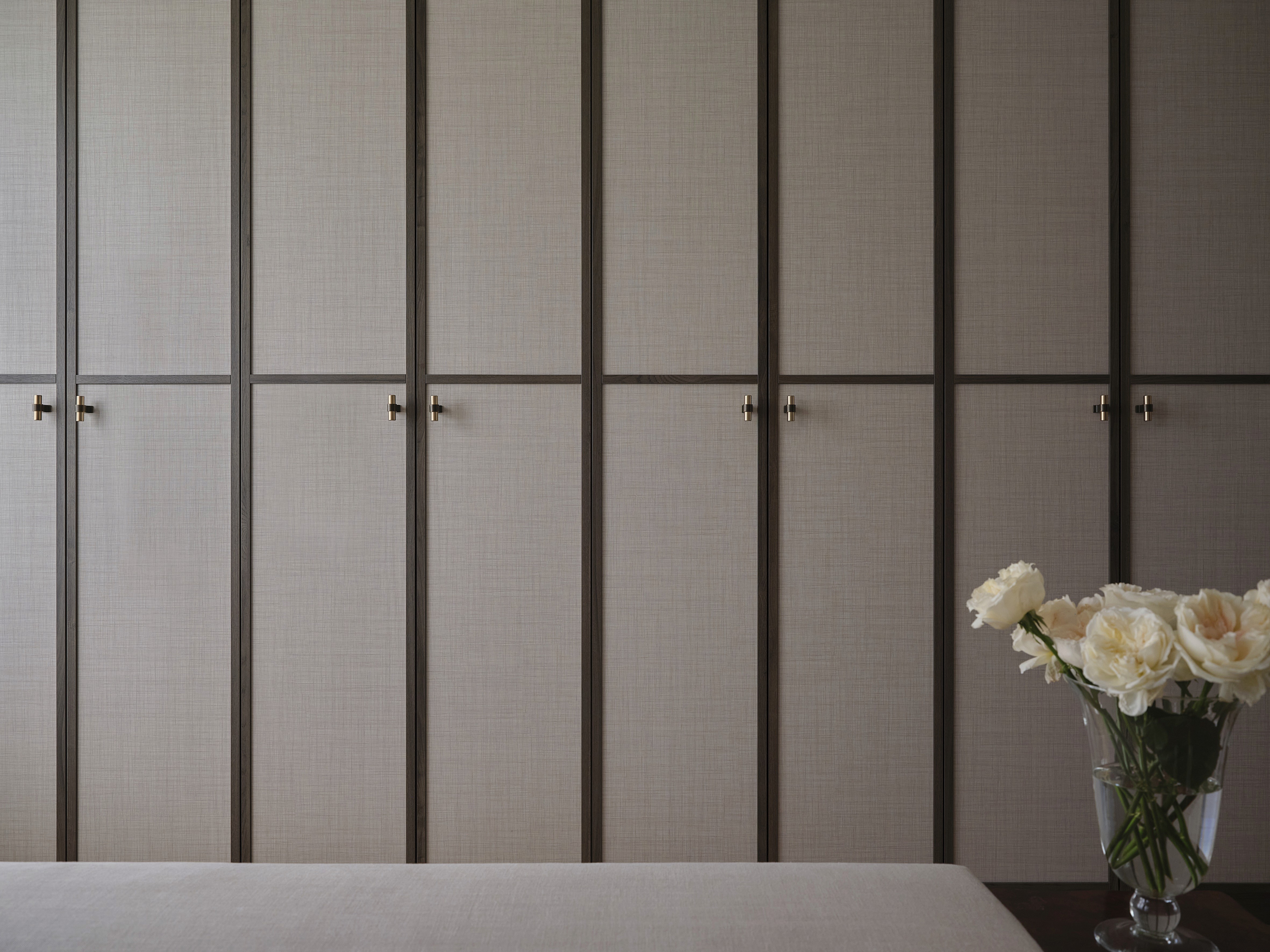 Floor-to-ceiling built-in wardrobes with linen-panelled doors, dark timber framing, brass T-bar handles, and fresh white roses in glass vase