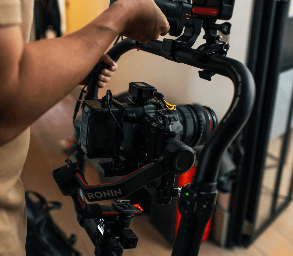 Een persoon houdt een professionele camera vast, gemonteerd op een Ronin-gimbalstabilisator, opgesteld binnenshuis, met een achtergrond met wazige apparatuur en zachte verlichting, wat de focus op videoproductieapparatuur benadrukt.