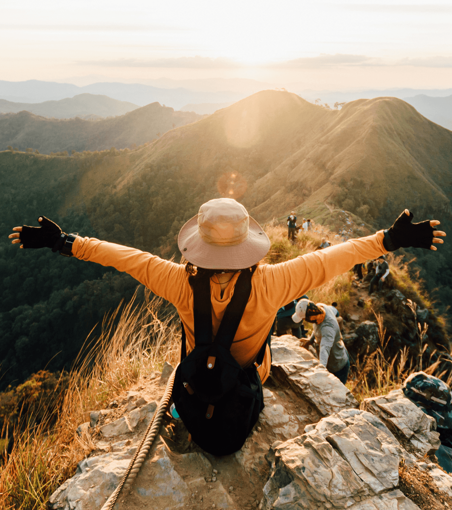 A hiker with arms outstretched stands on a mountain ridge at sunset.