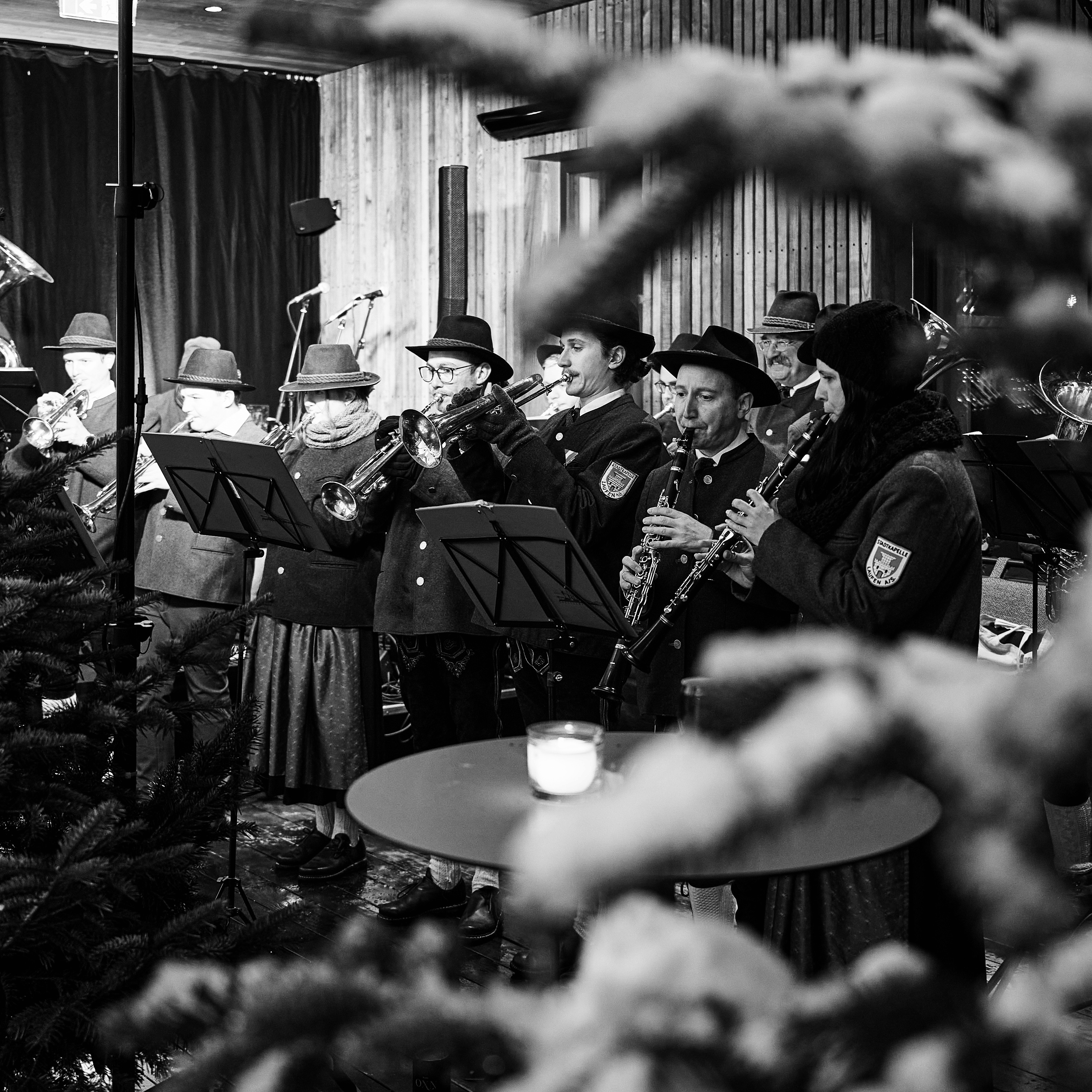 A monochrome image of a group of people gathered, possibly at a festive event, with a foreground of branches.