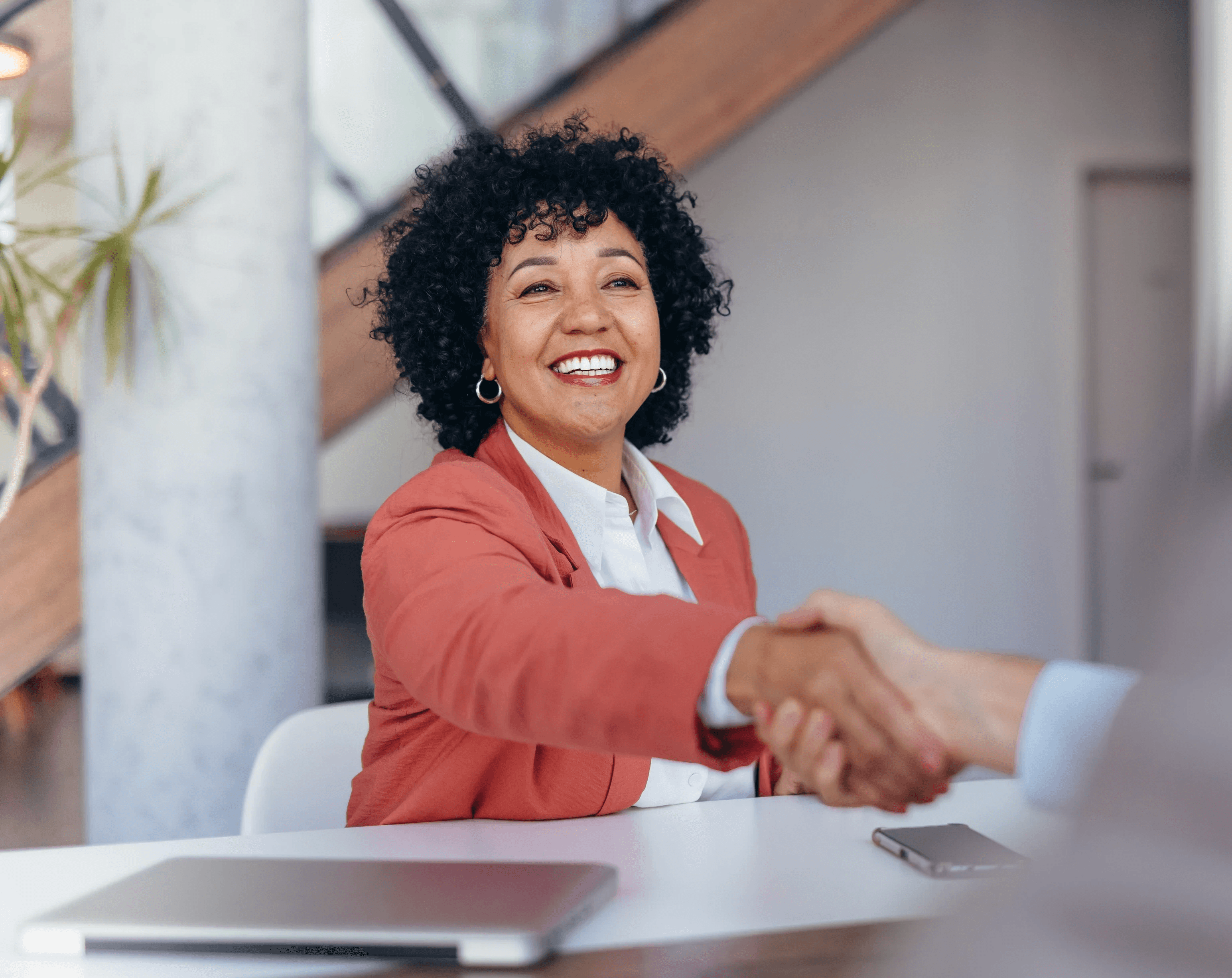 Smiling businesswoman shaking hands with someone across a table in an office setting.