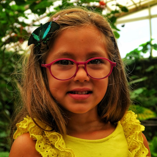 Chica joven con gafas y una mariposa en el pelo, vestida con un top amarillo, sonriendo en un entorno exterior lleno de hojas.