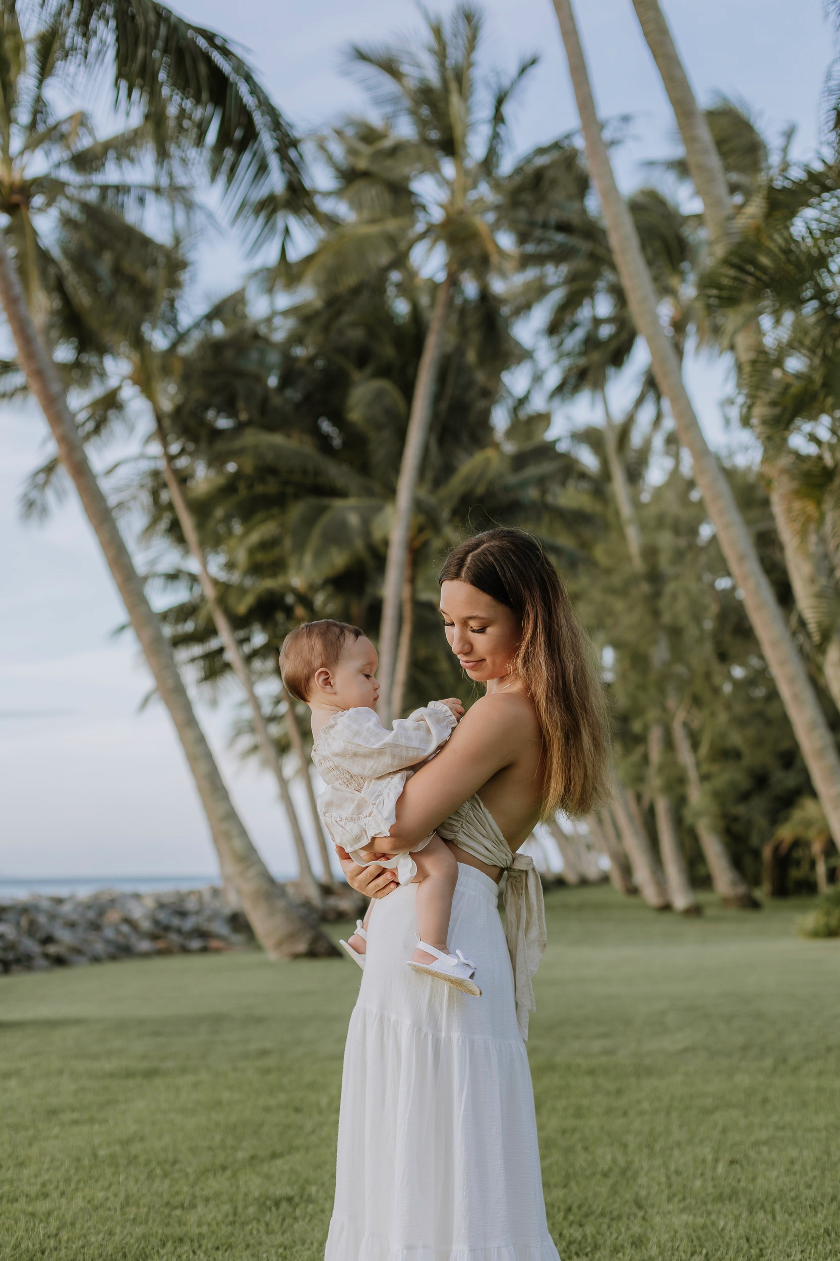 Mother cuddling her daughter while standing with palm trees in a coastal motherhood session