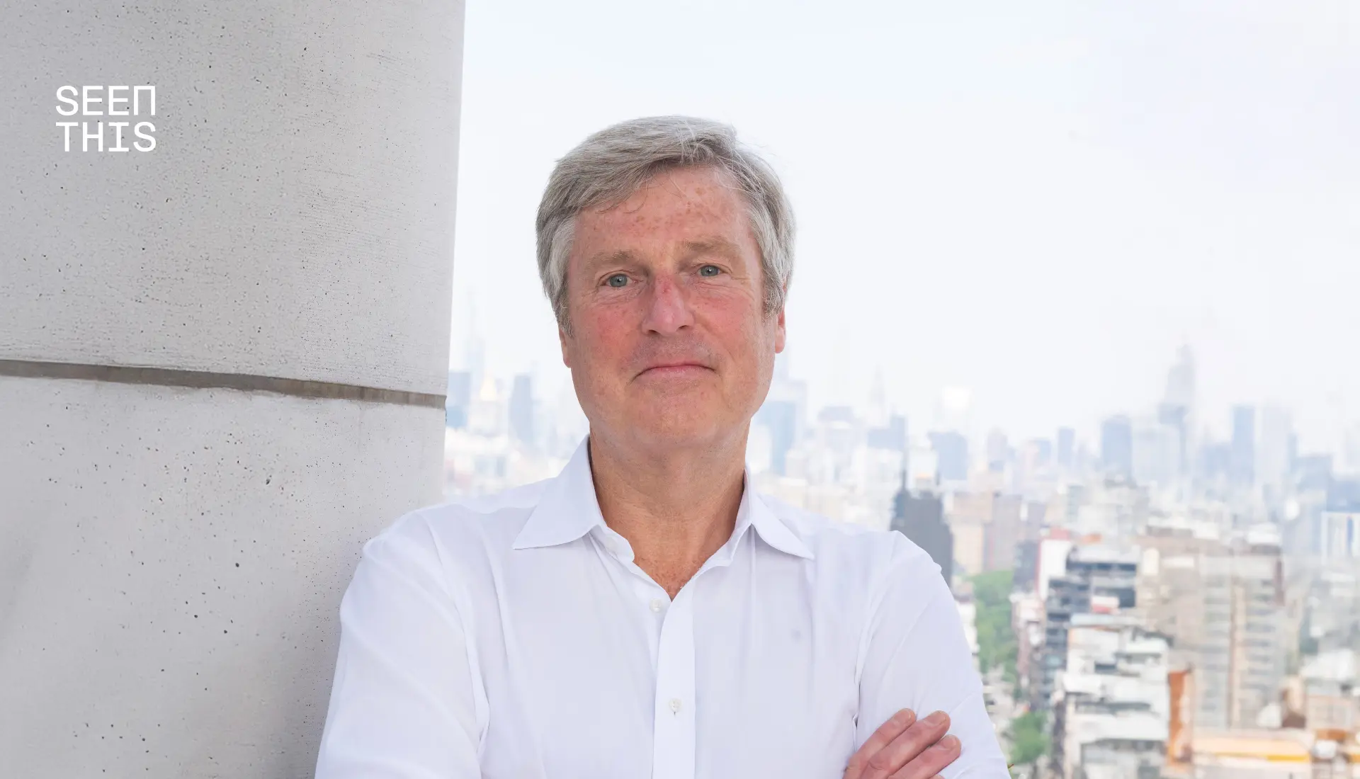 John Osborn, President US at SeenThis, posing in a white shirt with arms crossed against a blurred city skyline backdrop. The SeenThis logo is in the top left.
