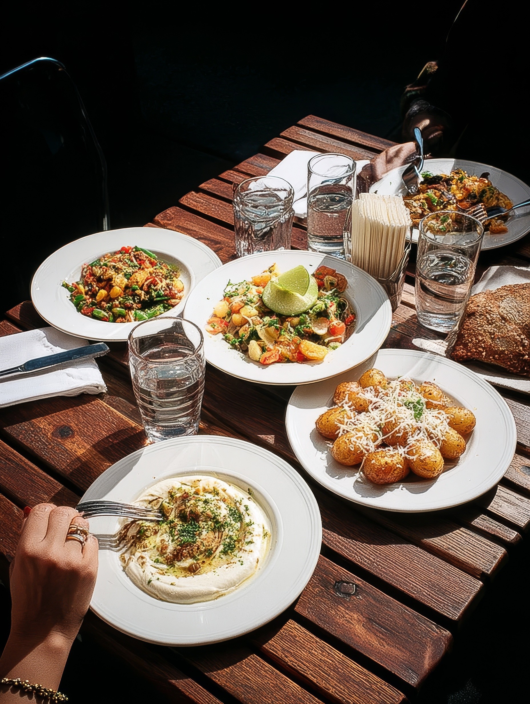 Wooden outdoor table set with multiple plates of food, including dips, salads, potatoes with cheese, and glasses of water, with hands reaching in to eat.