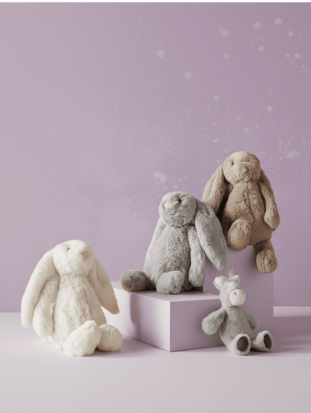 Four plush bunnies and a small donkey toy posed on pink blocks against a lilac background