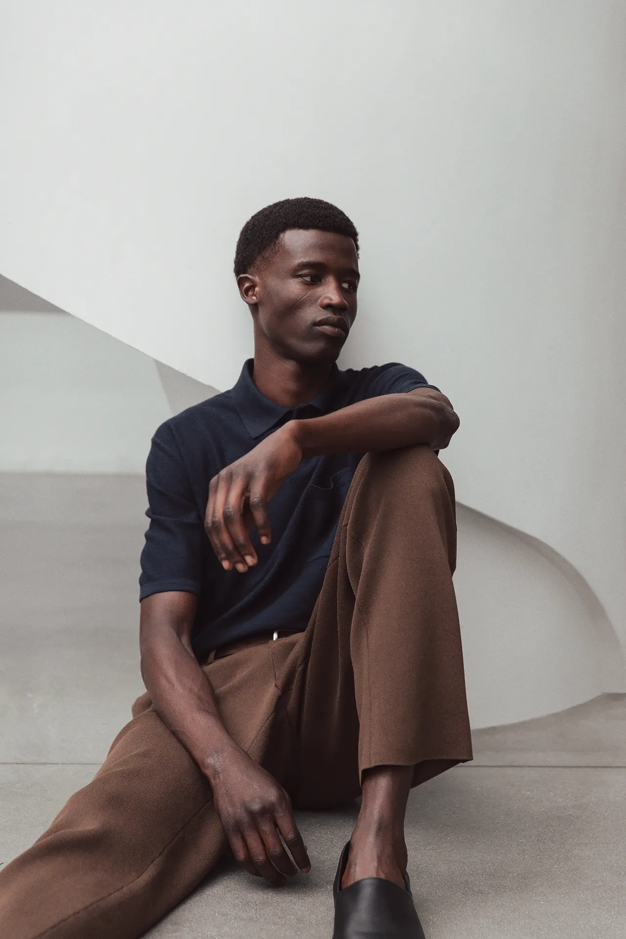 A male model seated on a white staircase wearing the Aida Barni SS26 collection men's navy blue short-sleeve knit polo shirt and brown trousers.