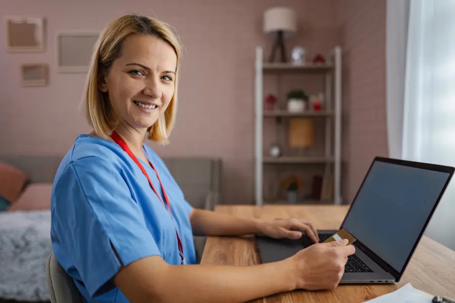 Smiling nurse in blue scrubs using AI hospital software on a laptop while holding a badge at a home desk.