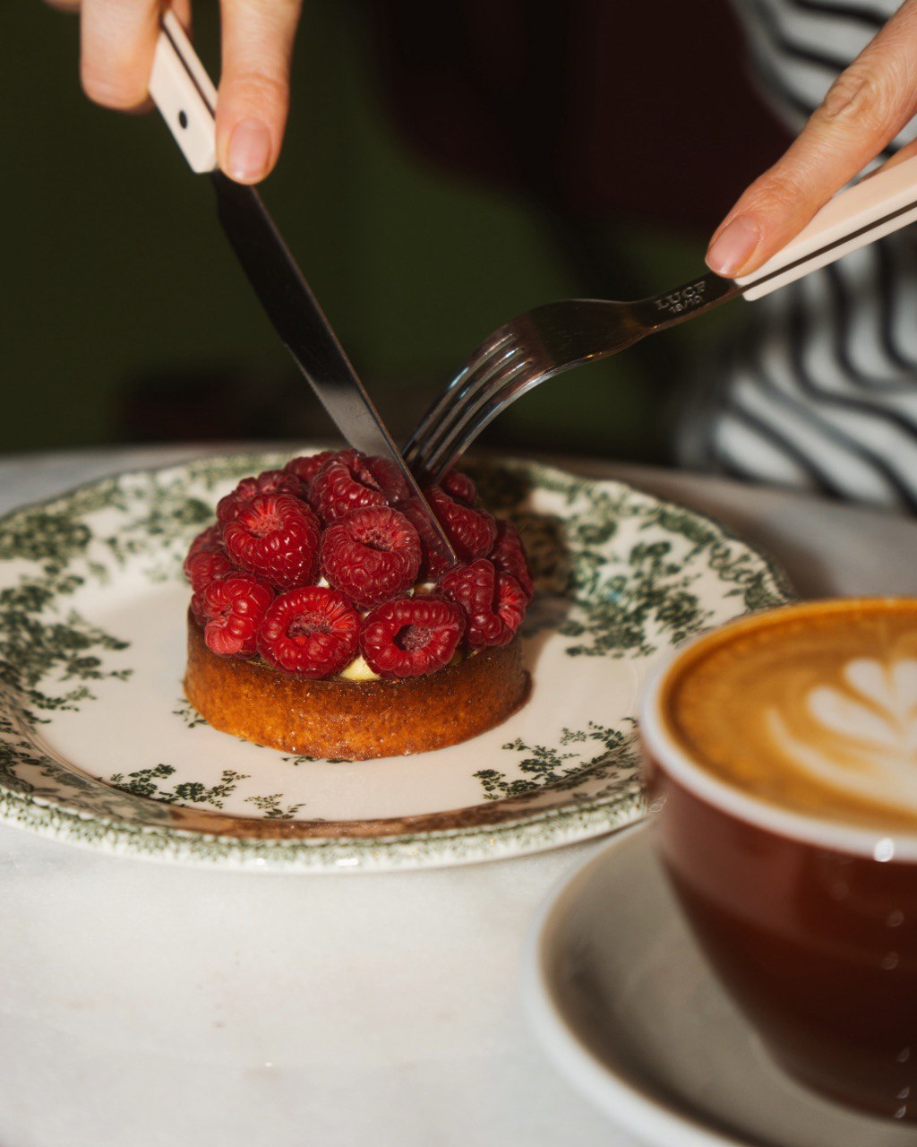 A close-up image of a person slicing through a raspberry-topped pastry on a decorative plate, accompanied by a cup of latte art coffee in a brown cup on a marble table.
