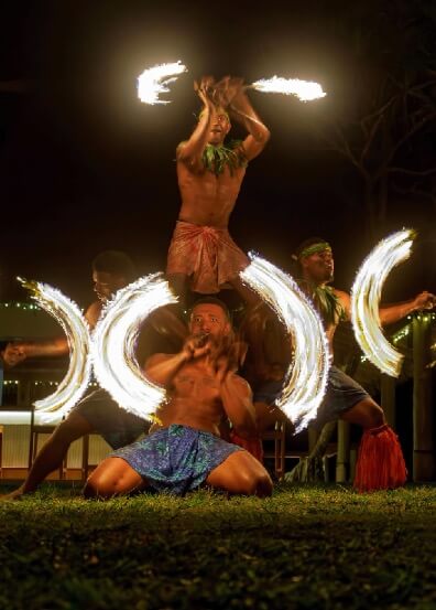 Fijian men perform a traditional fire dance at night, one balanced on shoulders, others creating fiery arcs.