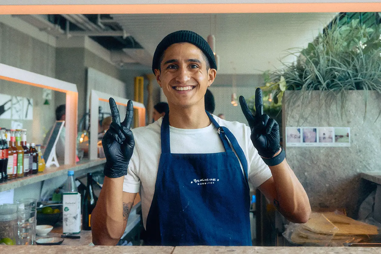 Chef smiling, wearing blue Benchside Burritos apron, holding up peace signs.