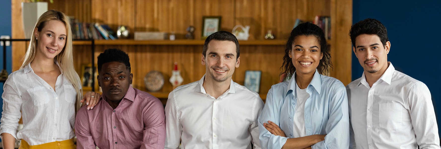A diverse group of five young professionals stand together, smiling and dressed in business casual attire, in a modern office with wooden shelving and blue walls.