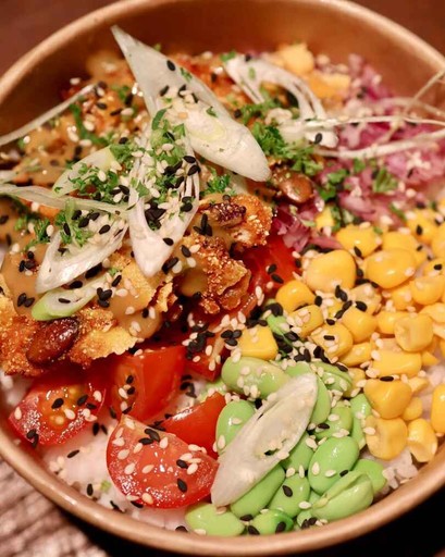 A colorful rice bowl topped with edamame, corn, tomatoes, onions, crispy chicken, sesame seeds, and sauce.