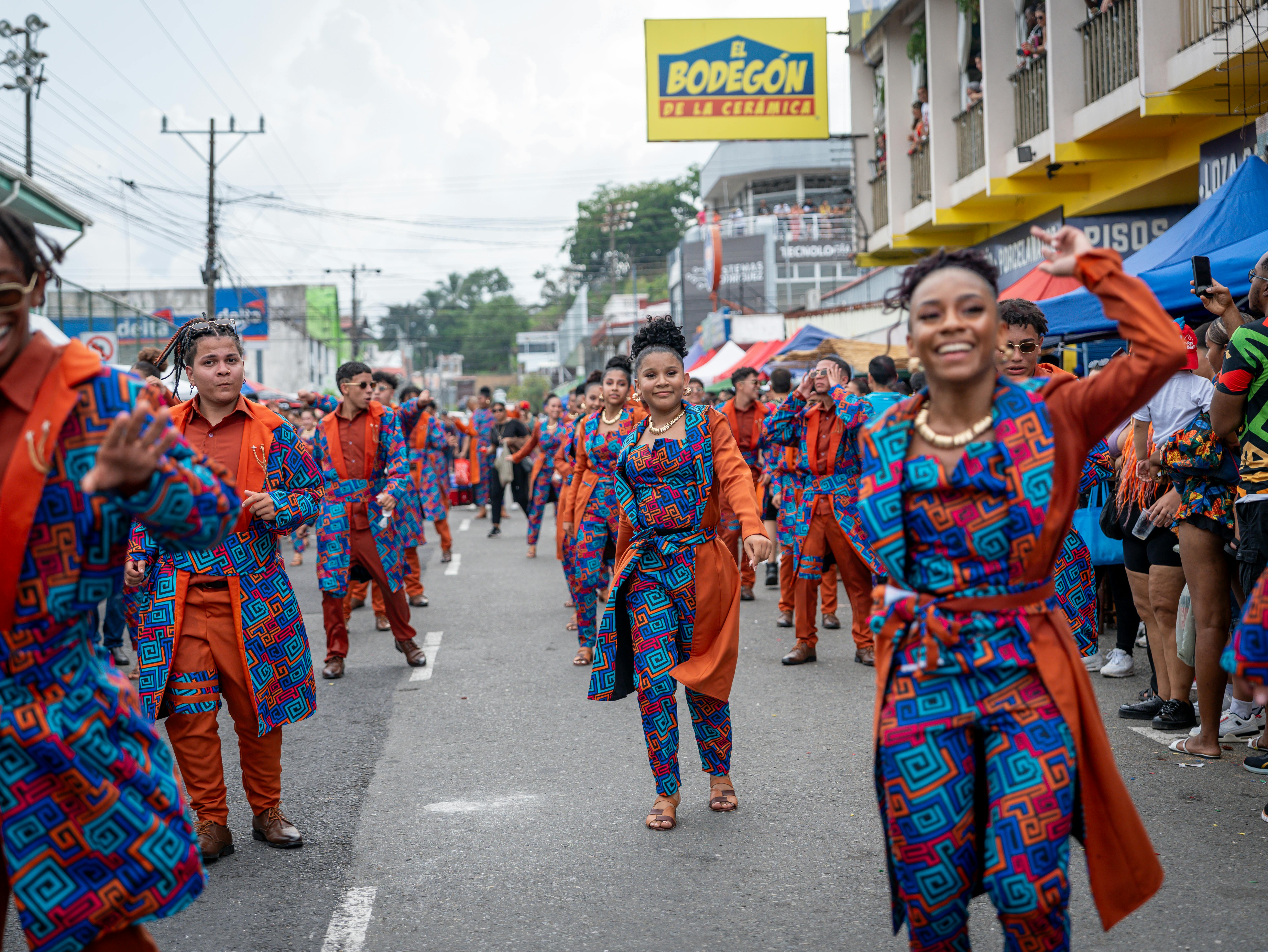 A lively street parade featuring a group of dancers in coordinated orange and blue geometric-patterned outfits. They are smiling and dancing down a paved road lined with spectators and commercial buildings.