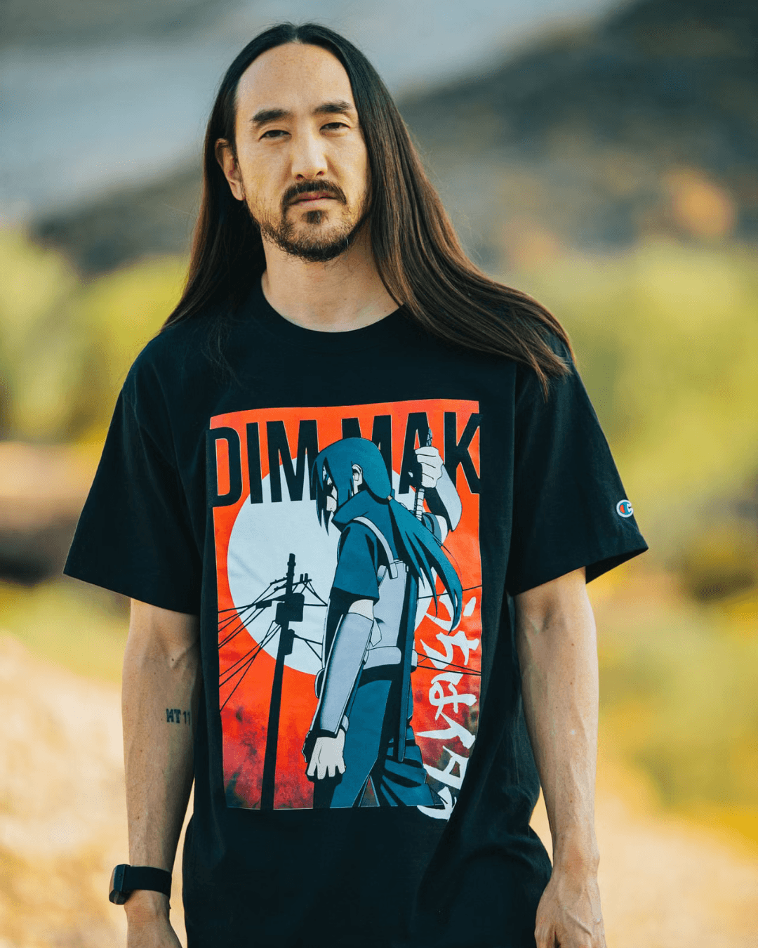 A person with long hair wearing a black graphic T-shirt stands in a natural outdoor setting.