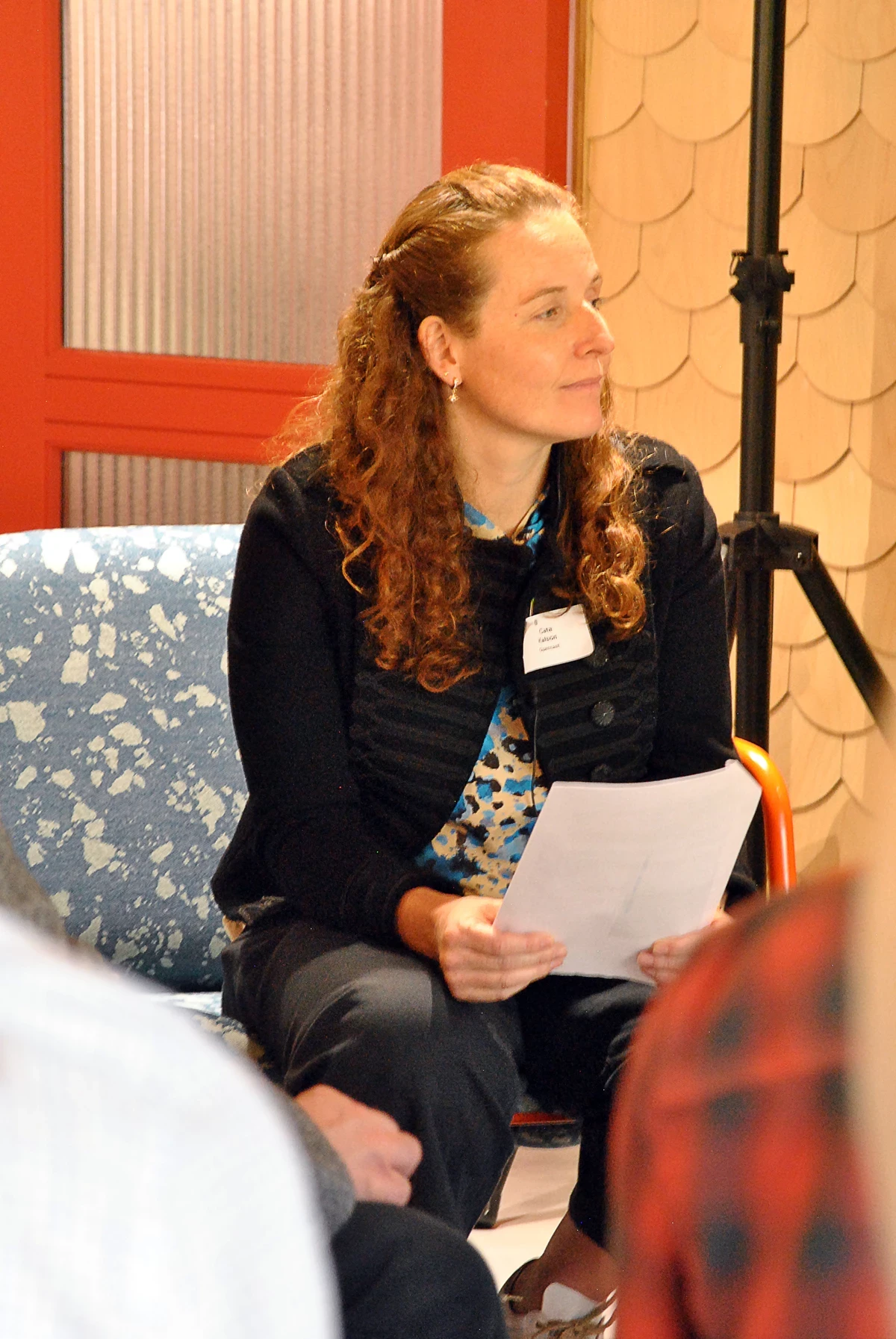 A woman in a black jacket sits on a chair holding a piece of paper