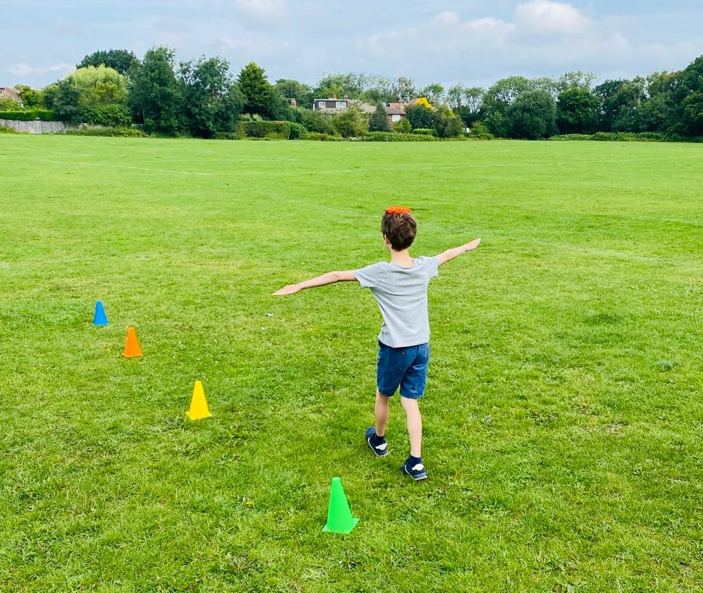 A child is walking in a field along a line of small cones as they balance a bean bag on their head. Their arms are outstretched to help them balance.