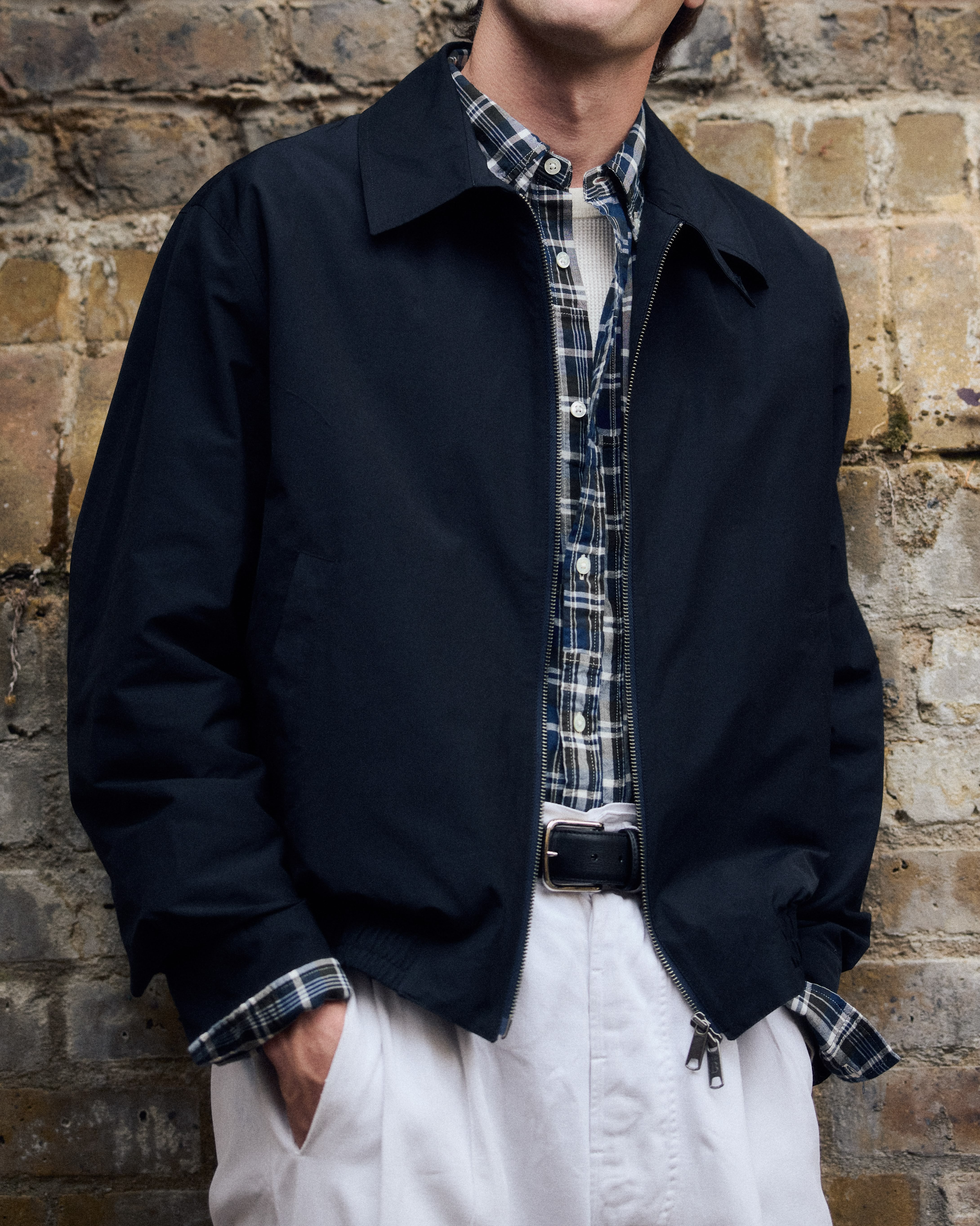 Le torse d'un mannequin devant un mur de pierre avec un blouson marine en drap de laine, pantalon blanc, chemise à carreaux