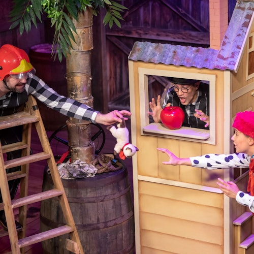 Three performers in a playful scene: one on a ladder, one in a window with an apple, and one reaching out with a red wig.