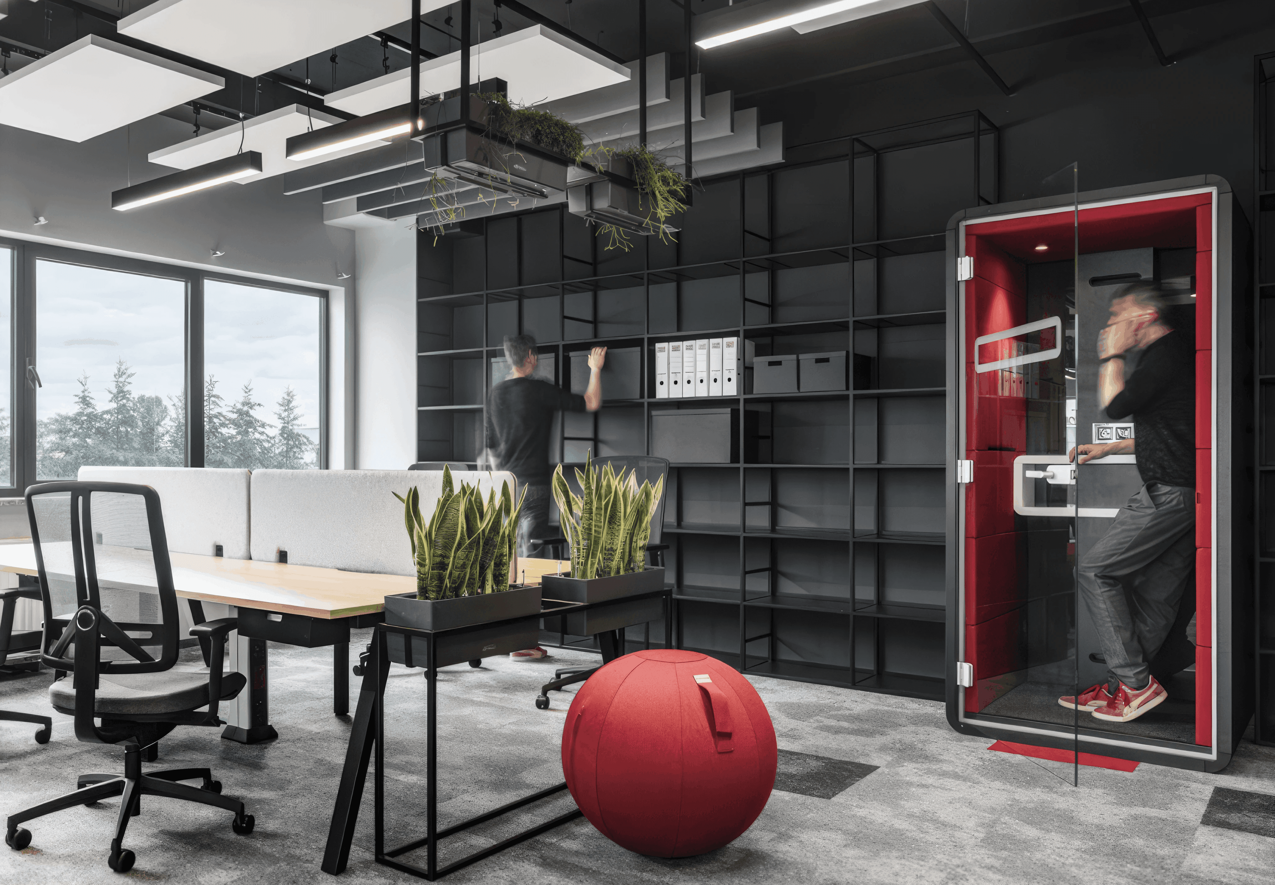 MW Interior - Modernes dunkles Büro mit Holzarbeitsplatz, schwarzem Regalrückwandsystem, rotem Sitzball, hängenden Pflanzen und roter Phone Booth" Beschreibung: Kraftvoller Büroraum in Anthrazit und Holz mit einer freistehenden Phone Booth in leuchtendem Rot, hängenden Deckenpflanzen und einem ergonomischen Sitzball – ein Büro, das Konzentration und Bewegung gleichermaßen fördert.