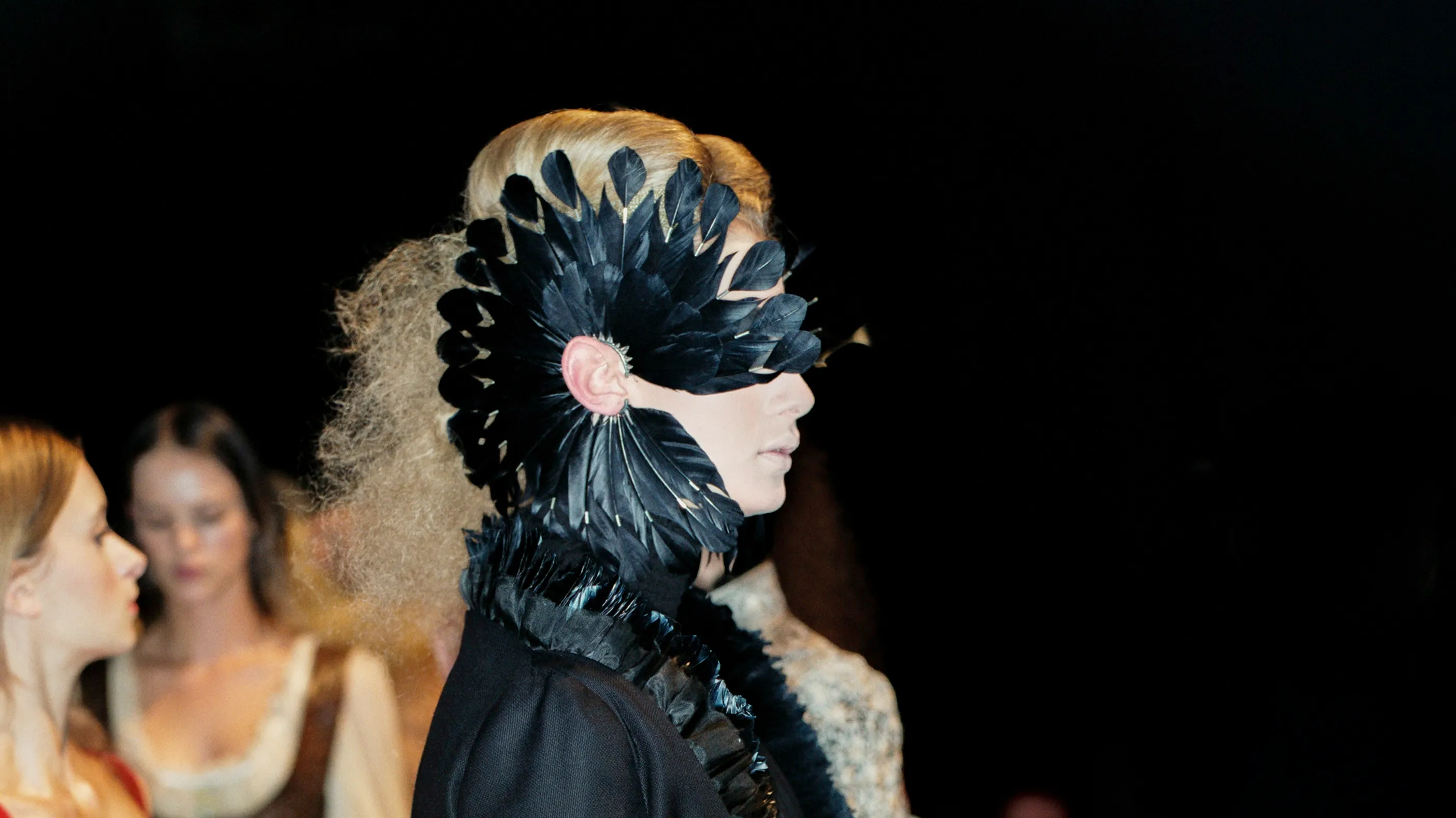 Profile view of a fashion model wearing a large, dramatic black feather ear cuff mask that frames the eye, designed by jeweller Shaun Leane.