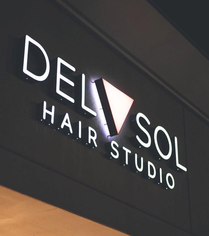 Illuminated Del Sol Hair Studio sign at night, featuring bold white lettering and the signature pink triangle glowing against a dark exterior.