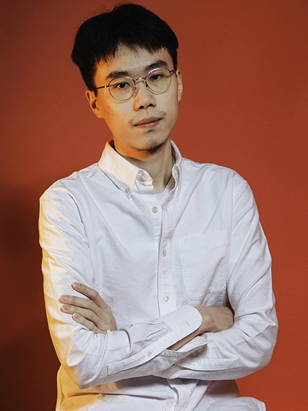 A young man with short black hair and round glasses stands against a solid orange-red background. He wears a white long-sleeved button-down shirt with his arms crossed over his chest. His expression is neutral, and he looks directly at the camera.