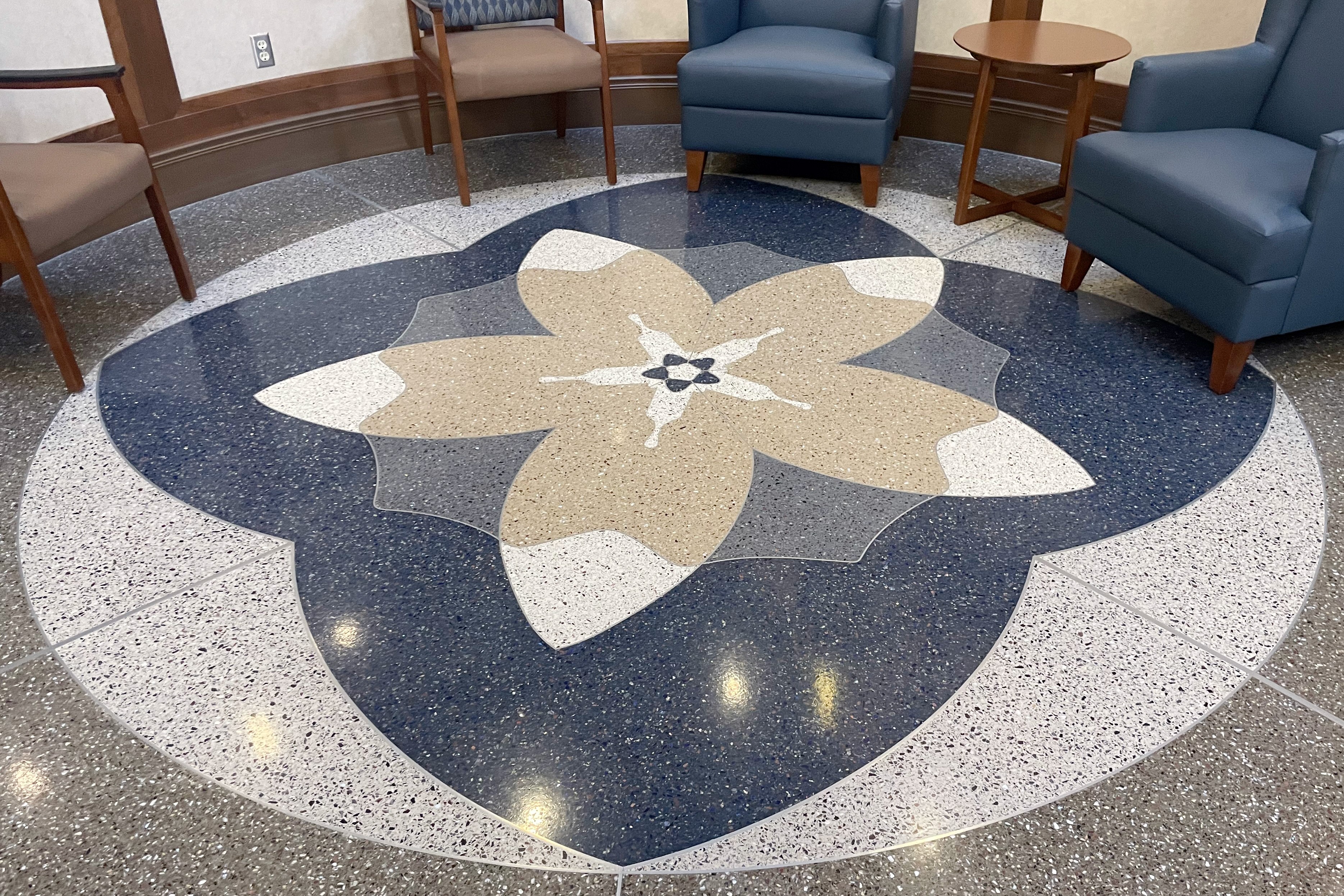 A close overhead view of the smaller second-floor terrazzo matchbrush medallion, its layered cream, tan, and blue petals radiating from a small star center, framed by a speckled aggregate border and waiting-area chairs at the Edith Sanford Breast Center.