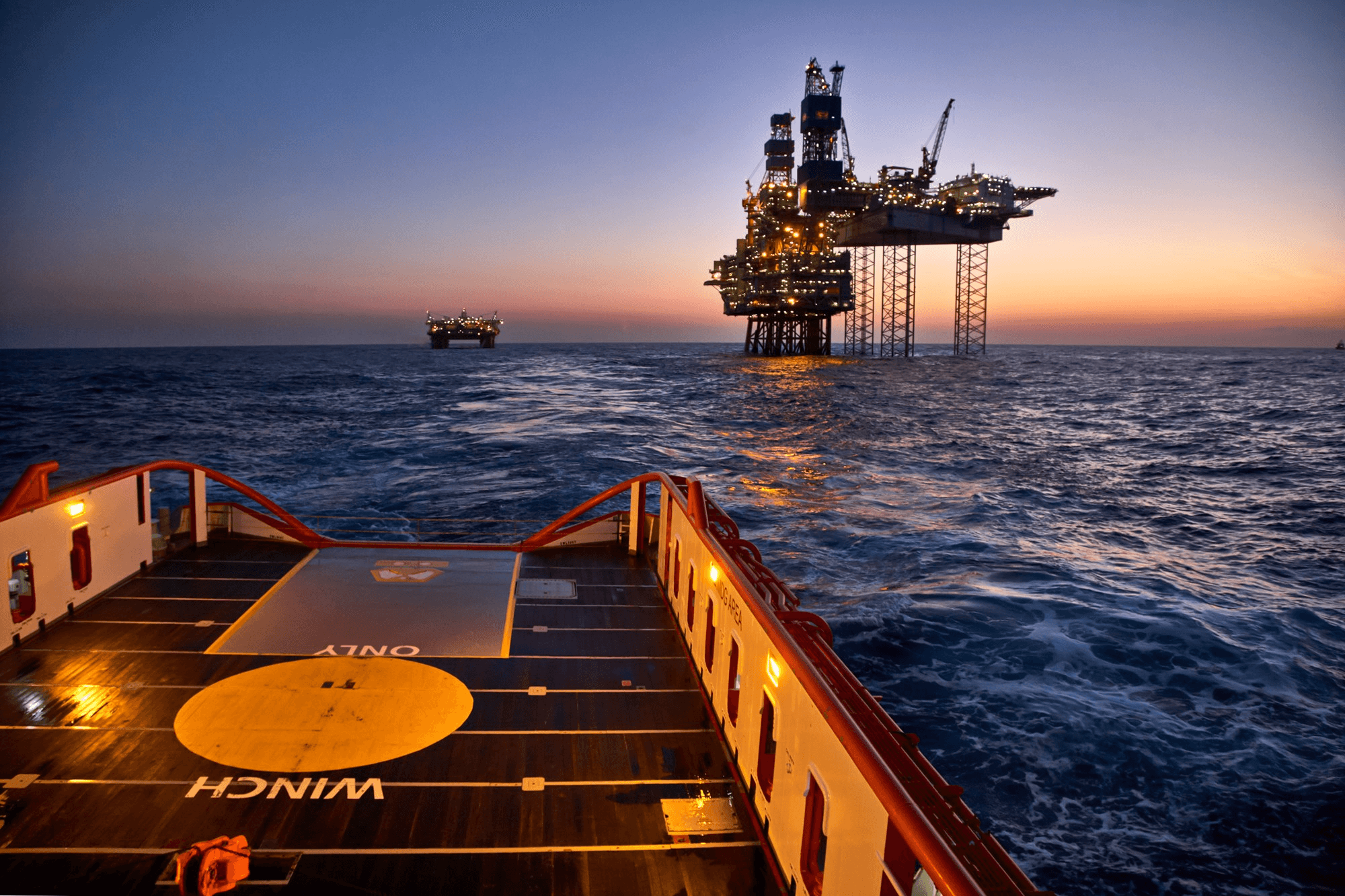 A ship approaches an offshore oil rig at sunset, creating a serene marine scene.