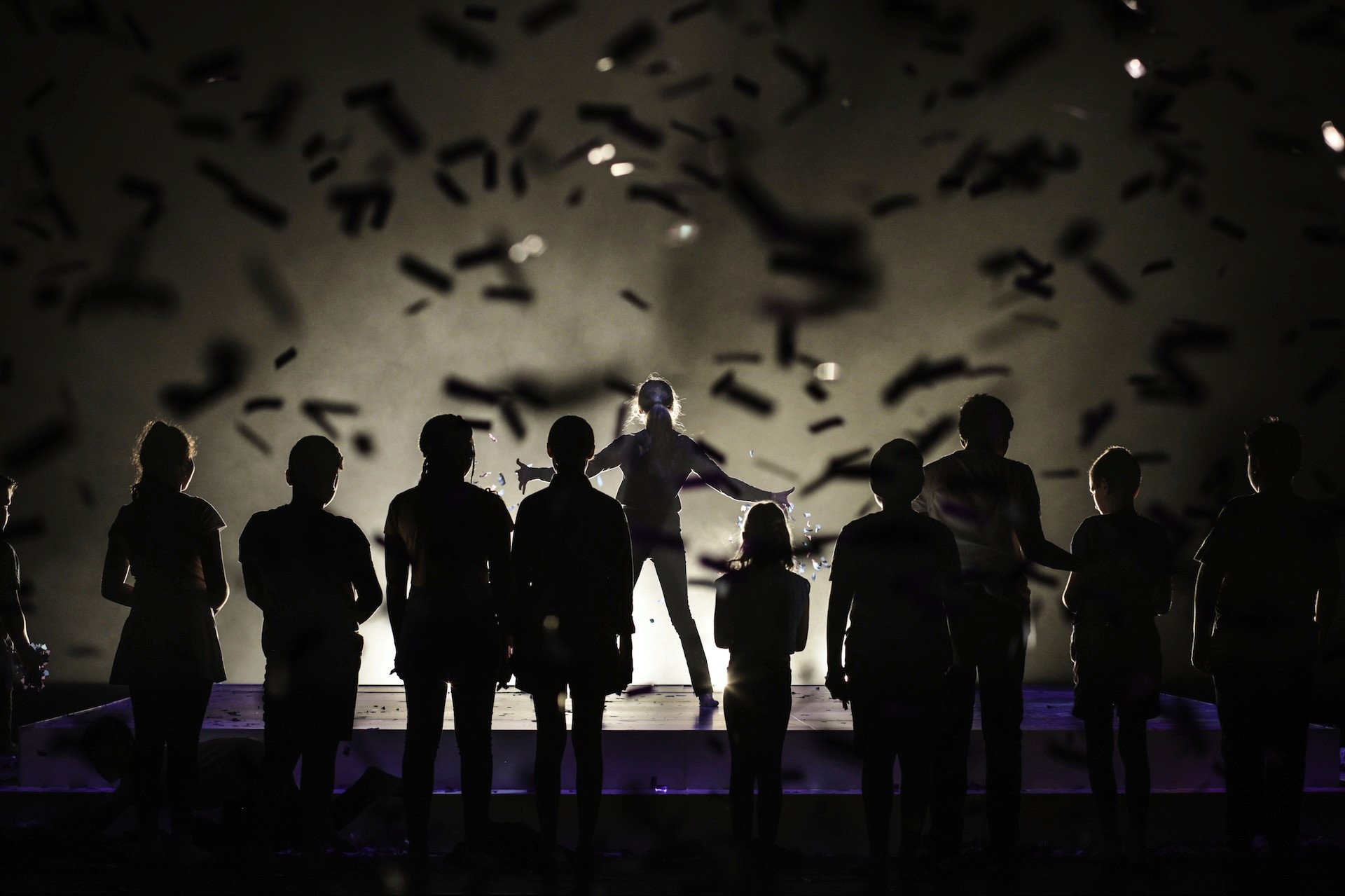 Silhouetted young performers face a bright spotlight as confetti rains across the stage. One performer stands elevated at the centre with arms outstretched, creating a powerful, triumphant image framed in falling shadows.